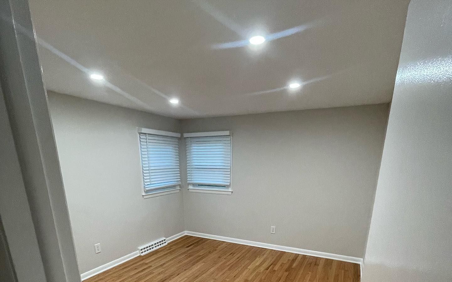 Empty room with recessed ceiling lights, two windows with blinds, and hardwood floors.