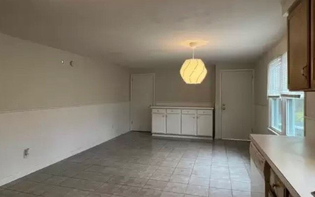 Empty room with white walls, cabinets, and a hanging light fixture. Gray tile floor and window.
