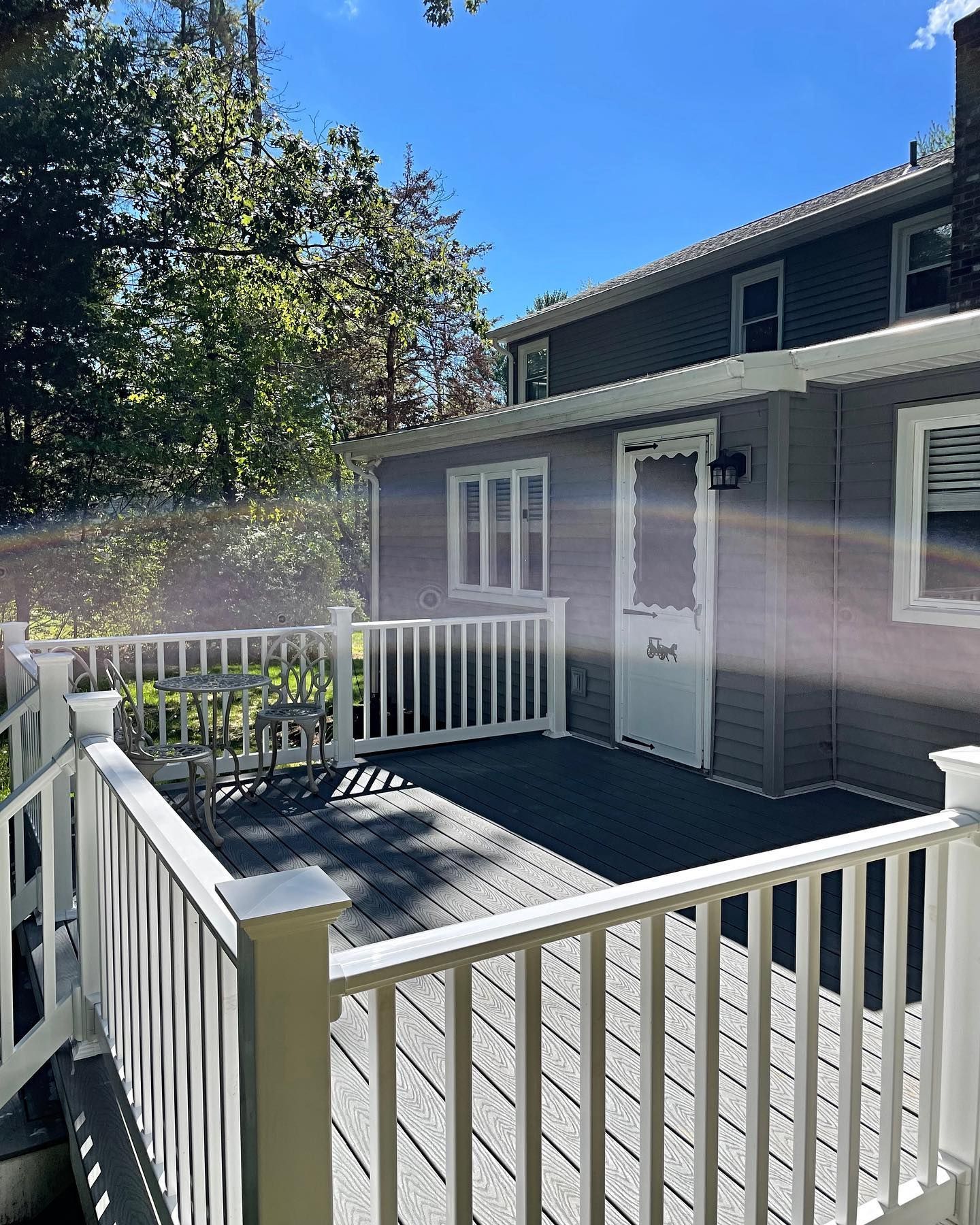 Wooden deck with white railing, gray siding building, trees in background, sunny day.