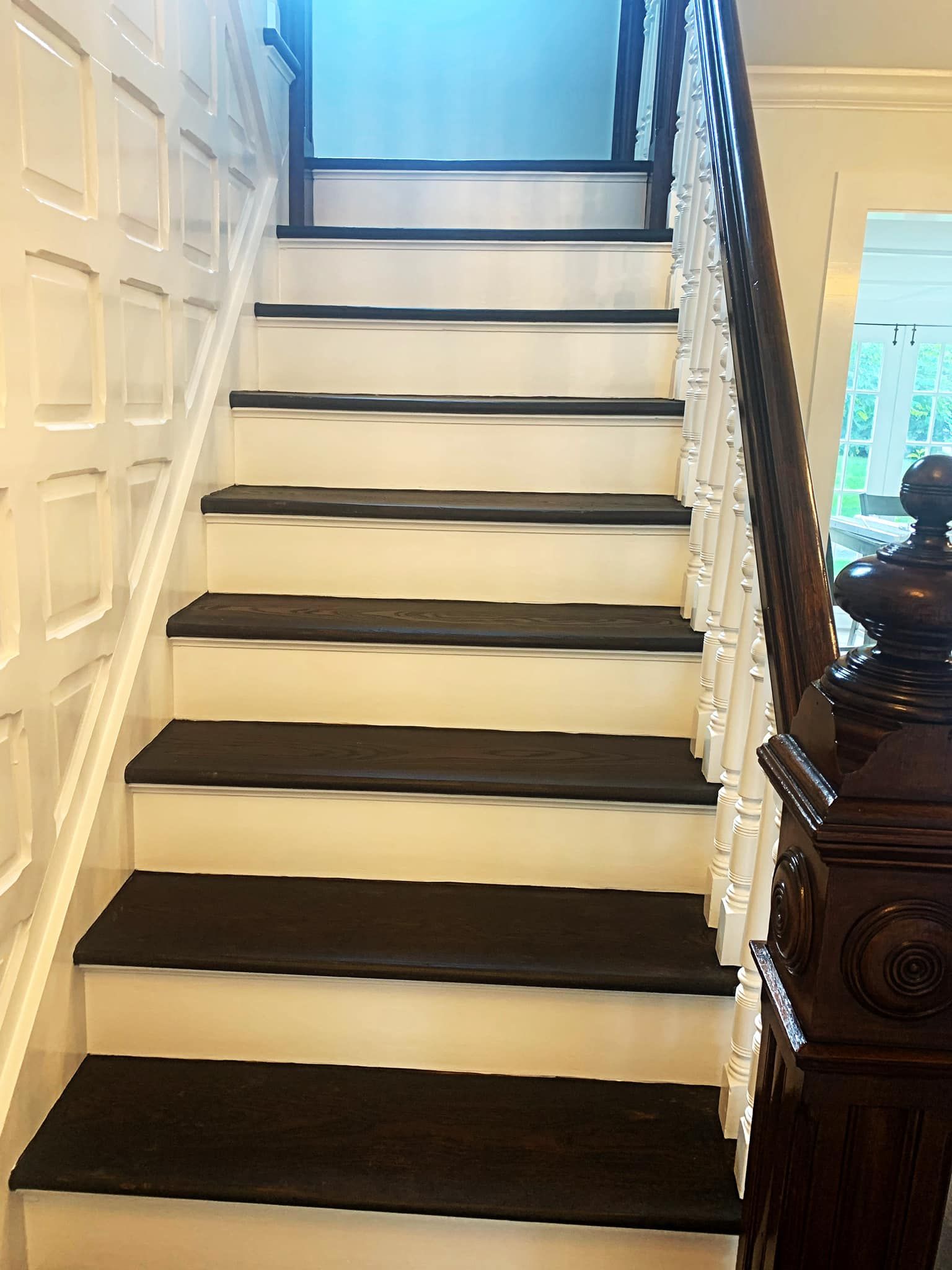 Staircase with dark treads and white risers, ornate dark wooden railing on right, white paneling on the left.
