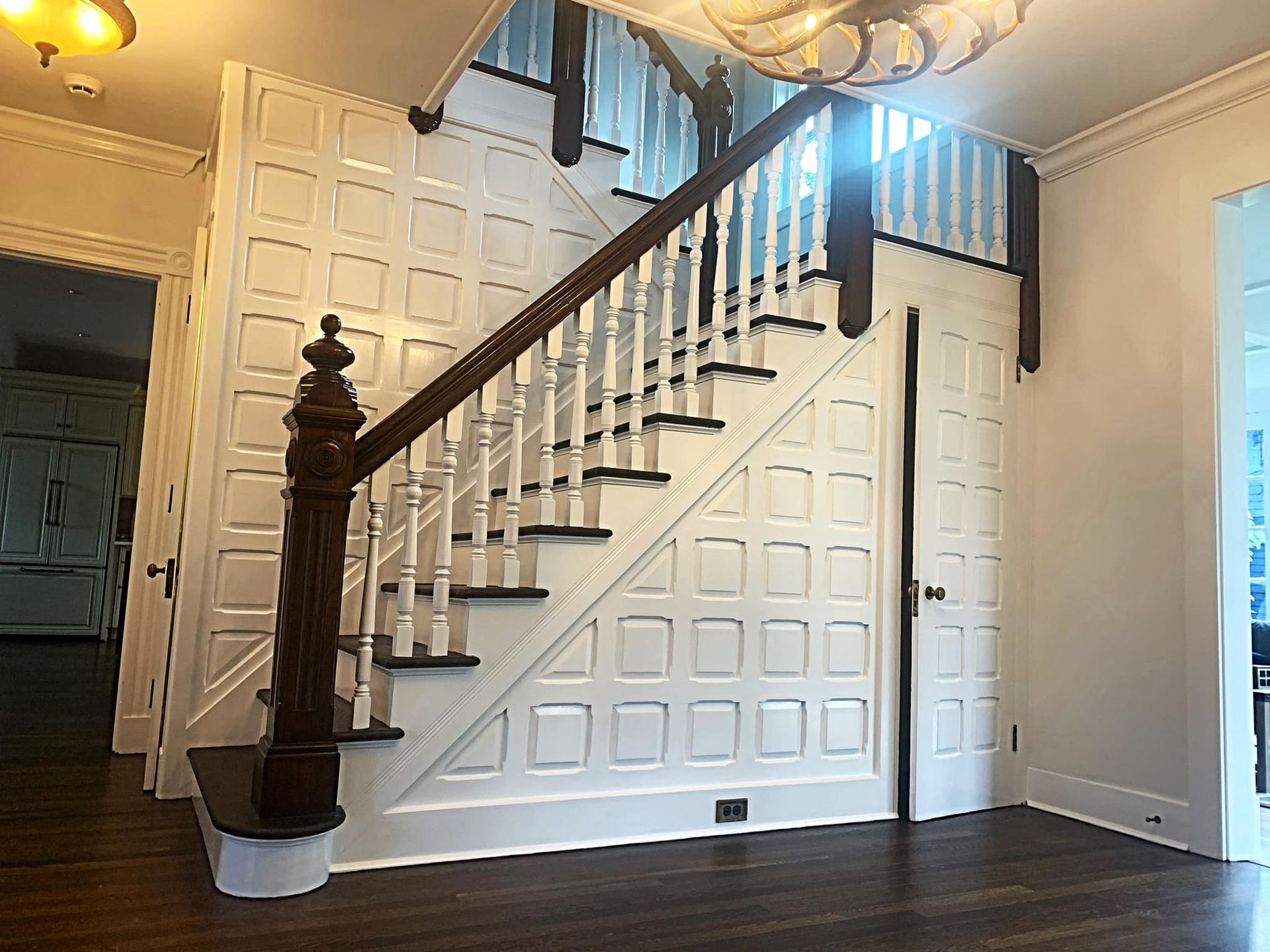 Staircase with white paneled walls, dark wood banister and steps, and a large light fixture.
