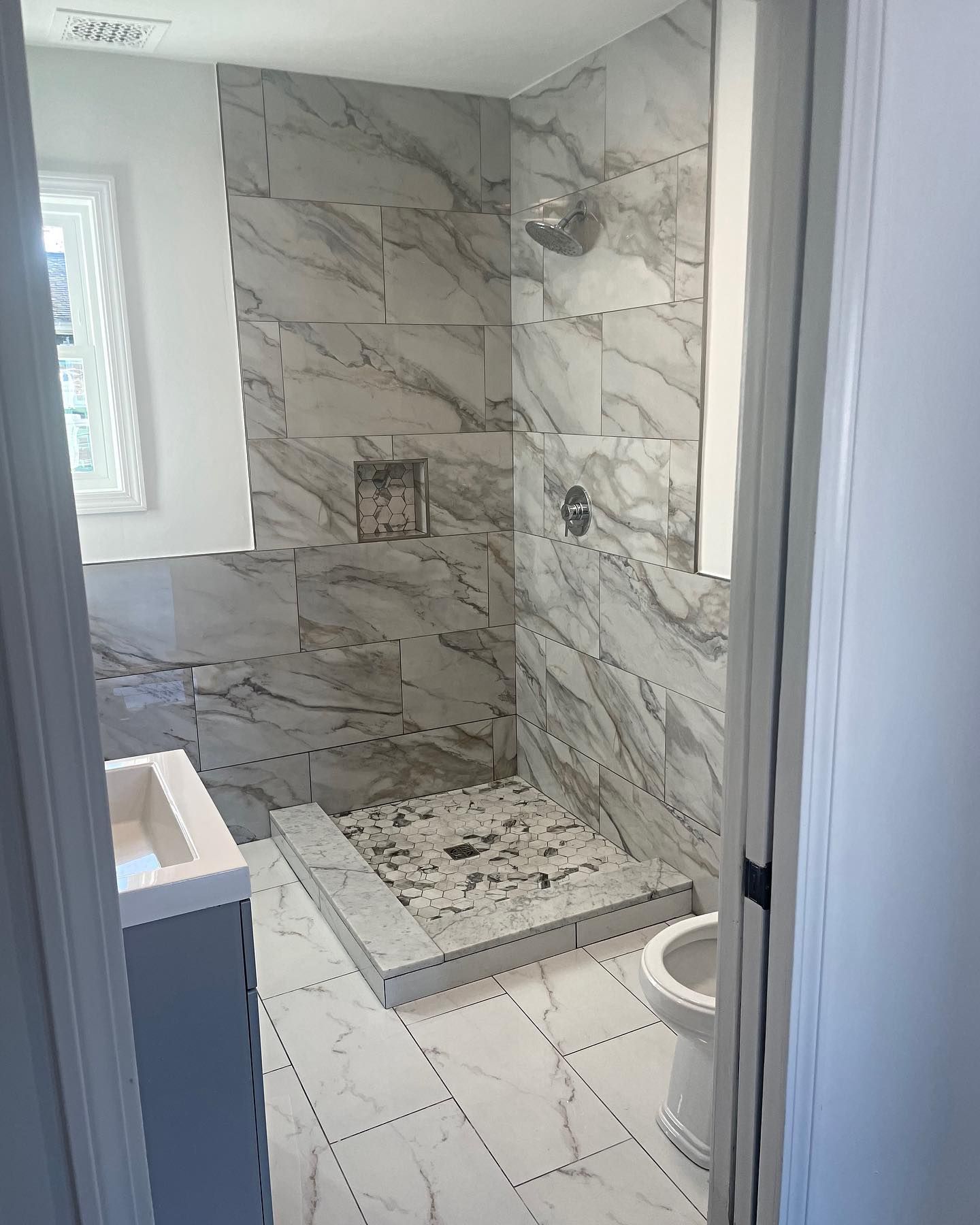 Bathroom with marble-look tile walls and floor, shower, toilet, and a window.