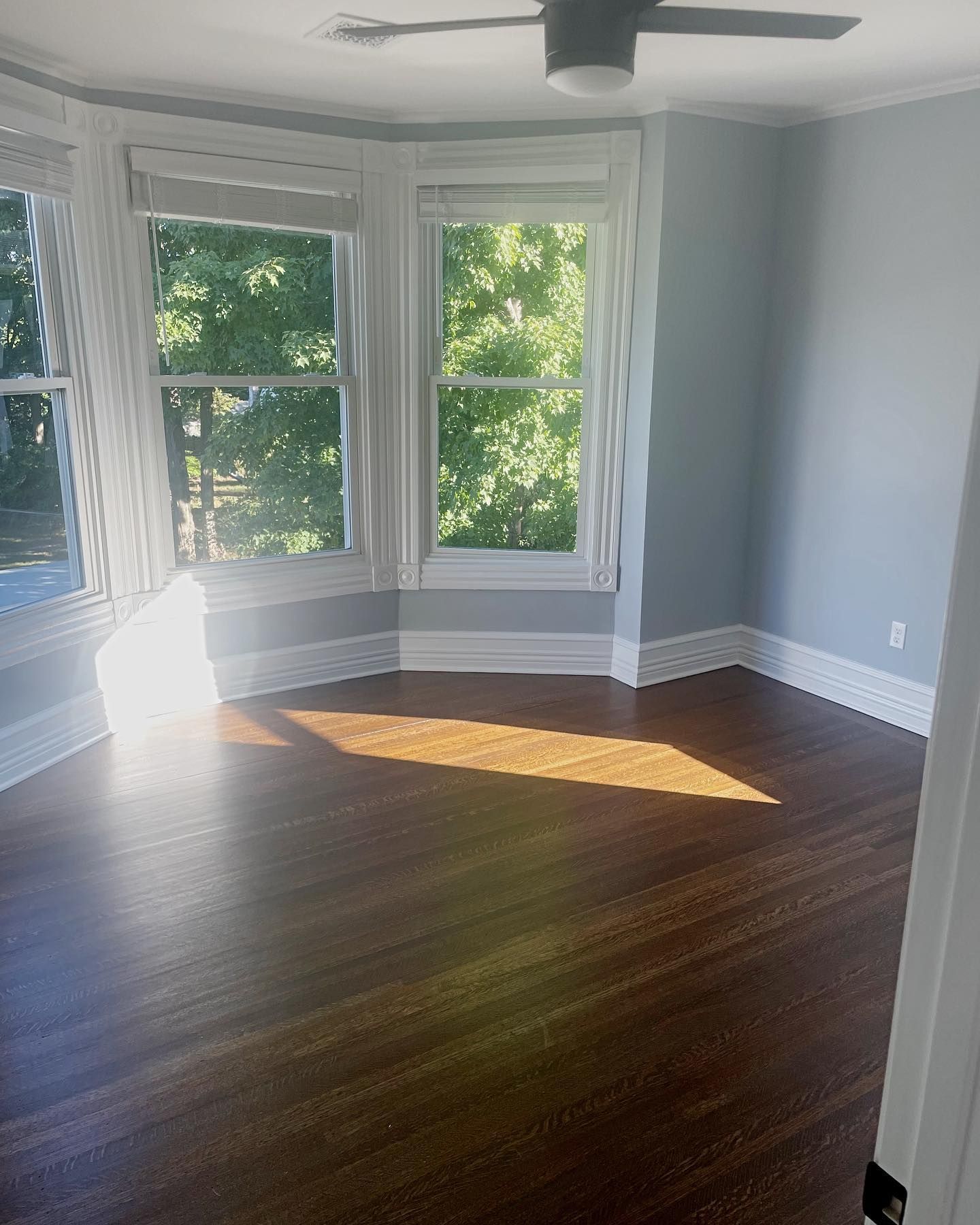 Empty room with a bay window, hardwood floor, and a ceiling fan. Walls are light blue.