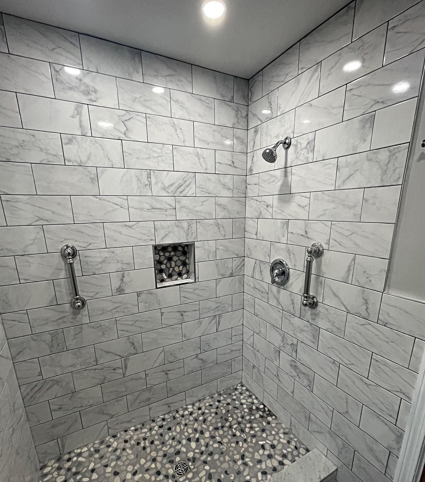 Shower with white marble-like tile walls and pebble floor. Includes shower heads, grab bars, and recessed shelf.