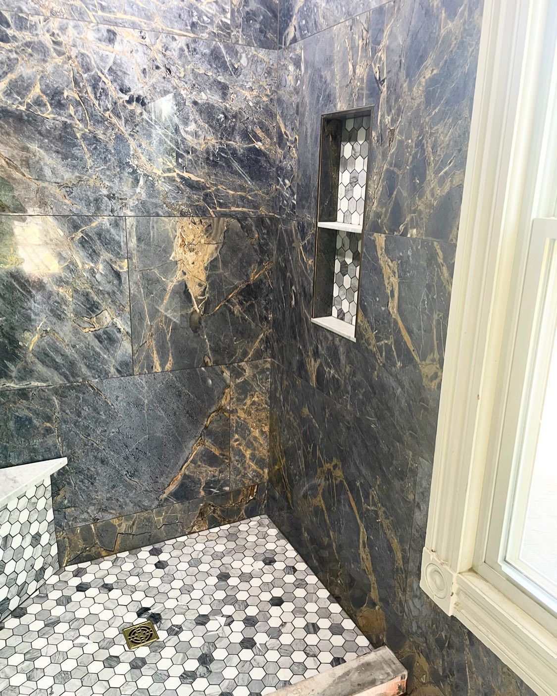 Shower with black and gold marbled tile, pebble floor, and built-in shelves next to a window.