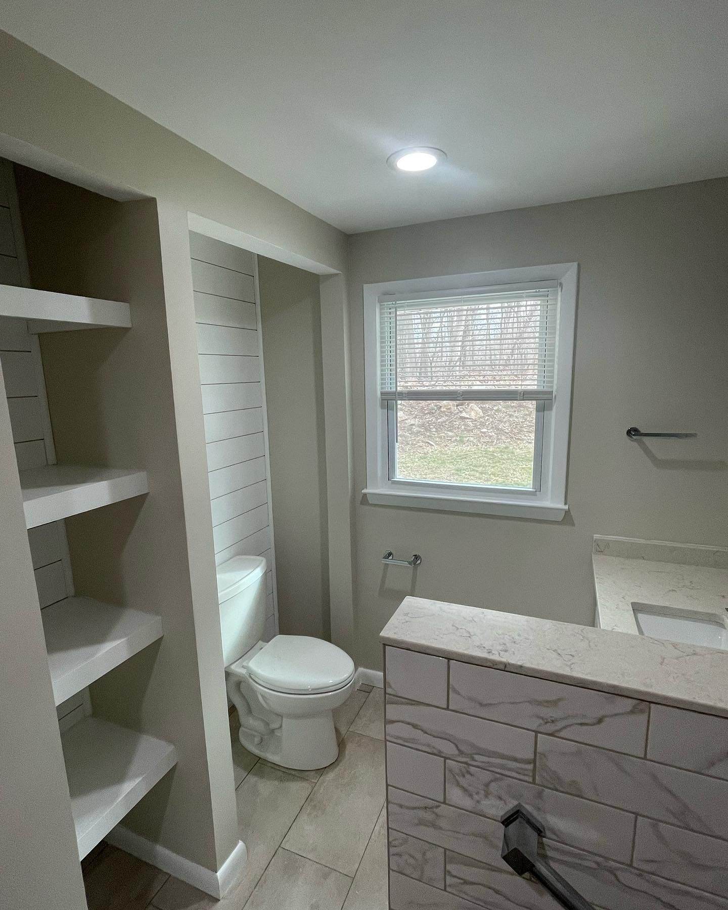 Bathroom with toilet, built-in shelves, window with blinds, and tiled countertop.