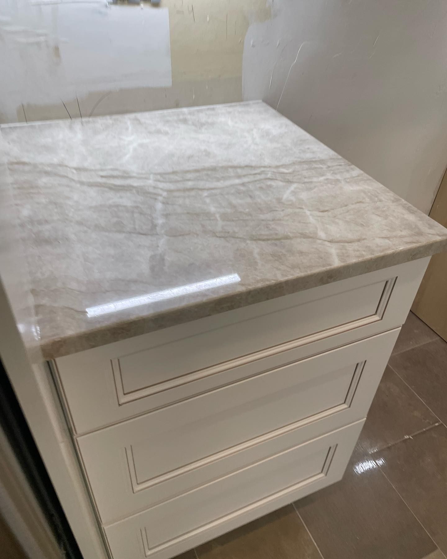 Cream-colored cabinet with a light brown marble countertop. Three drawers with inset panels. Light and shadow present.