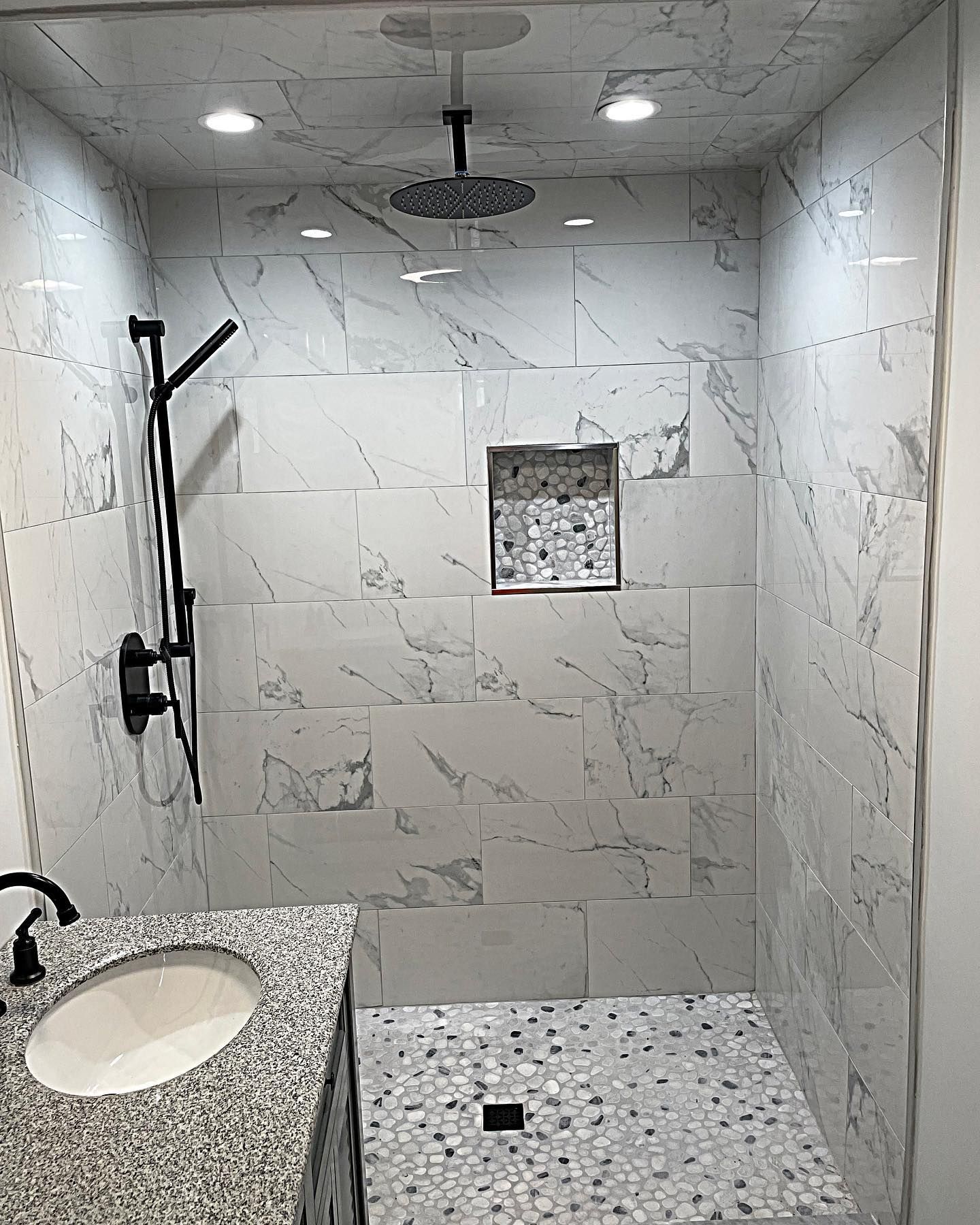 Modern bathroom with marble tile shower, black fixtures, pebble floor, and granite countertop.