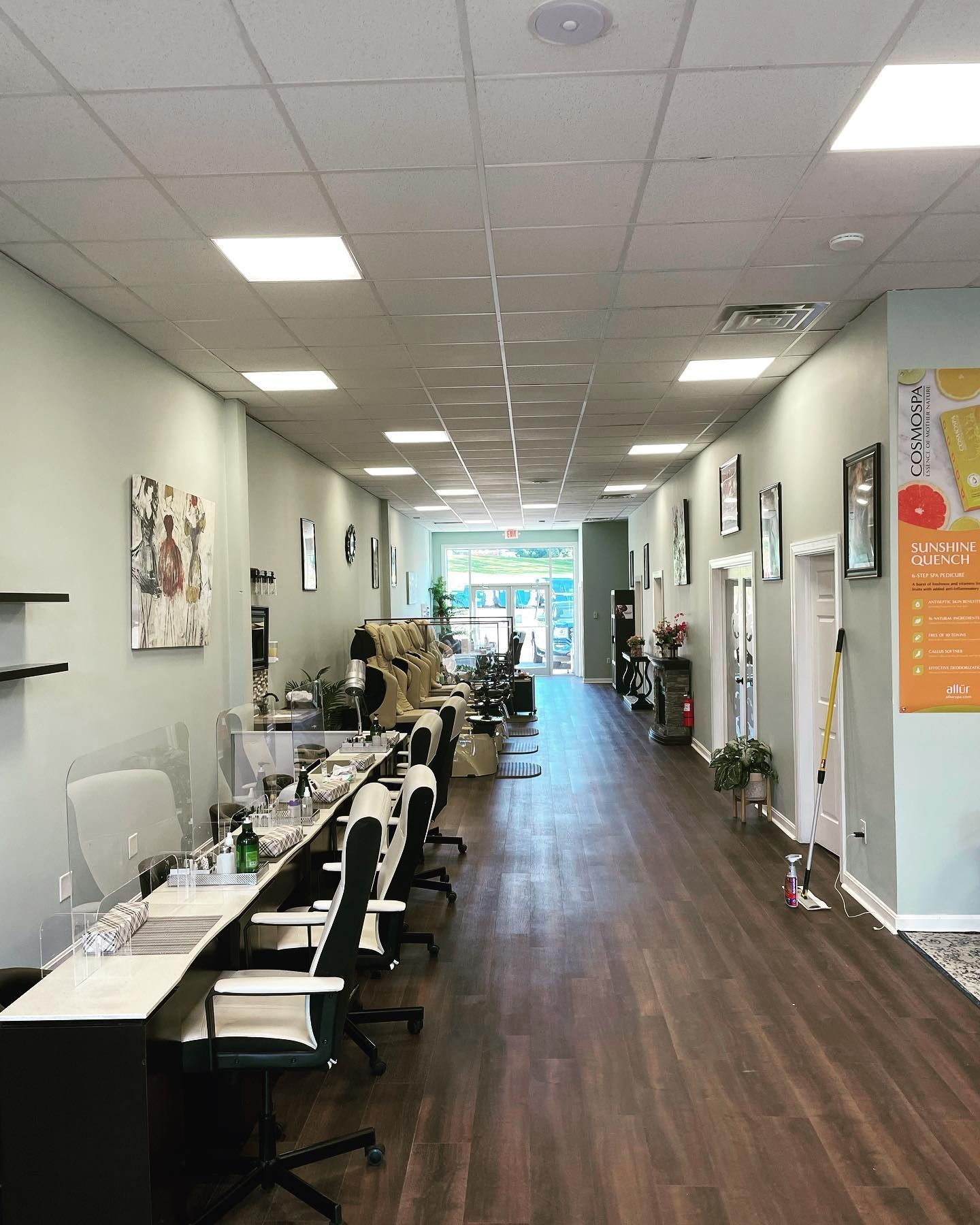 Nail salon interior with rows of manicure stations, chairs, and dark wood floors. A door at the end opens outside.