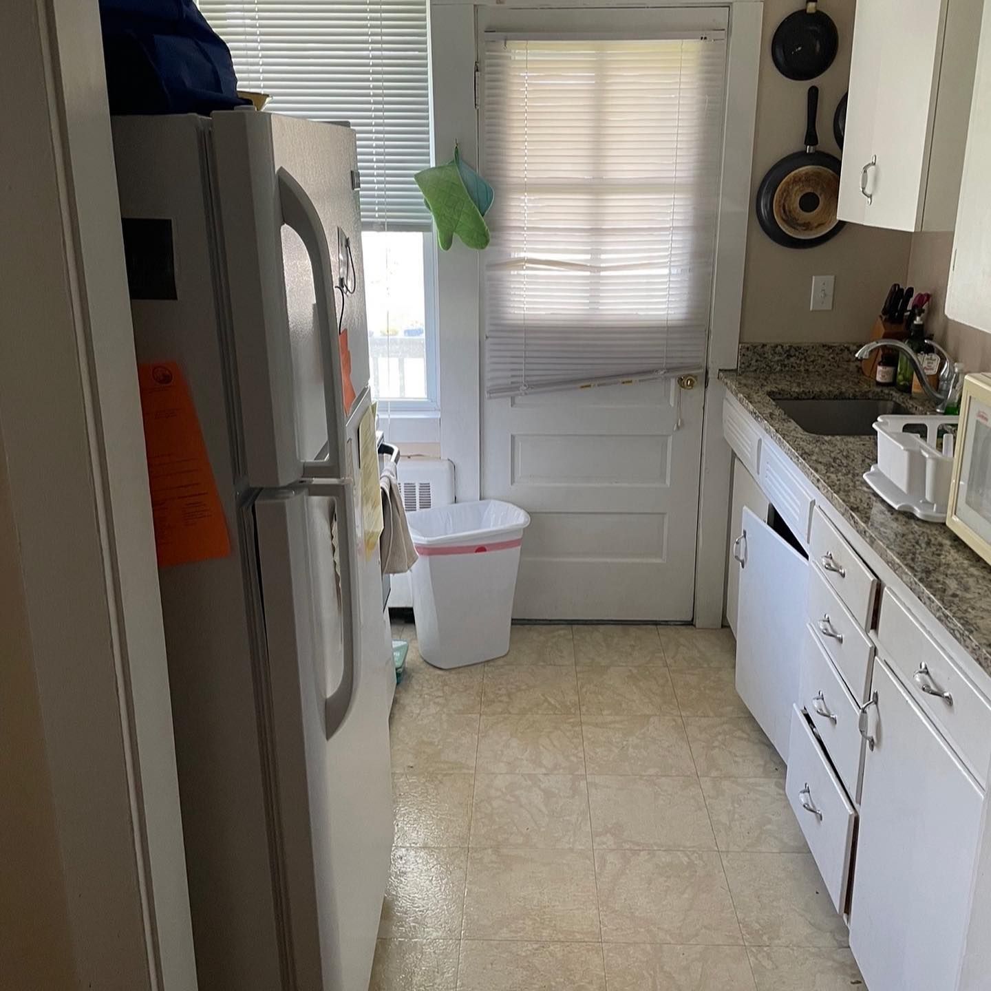 Small kitchen with white appliances and cabinets, window, door, and a trash can.