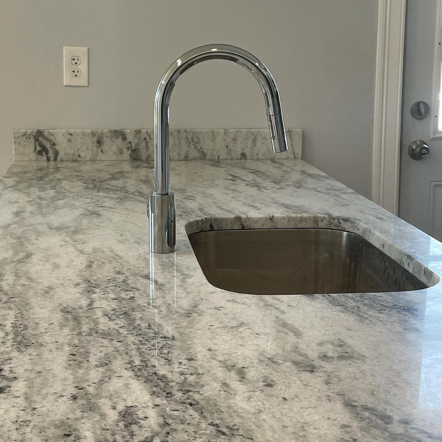 Kitchen counter with a faucet and sink, made of granite. A light-colored wall and door are in the background.