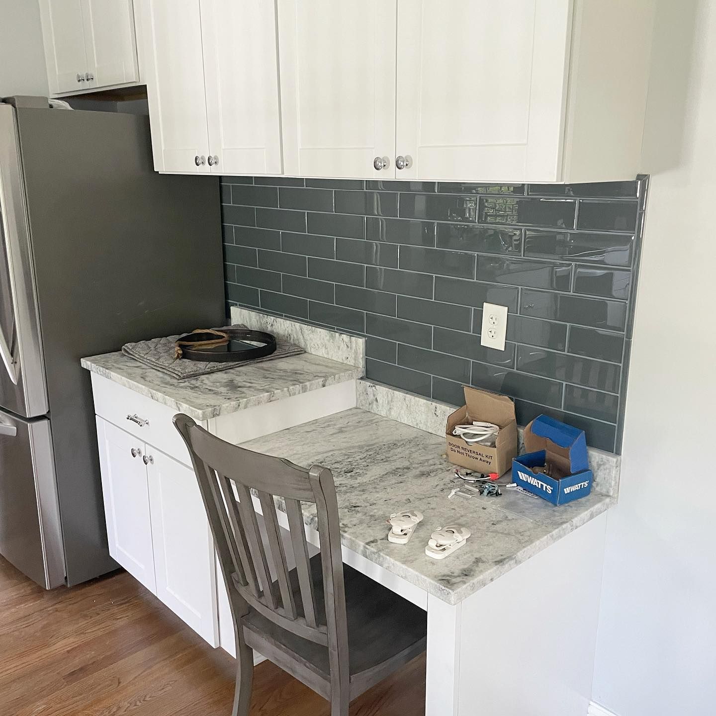 Kitchen counter with built-in desk, white cabinets, grey tile backsplash, silver fridge, and grey chair.