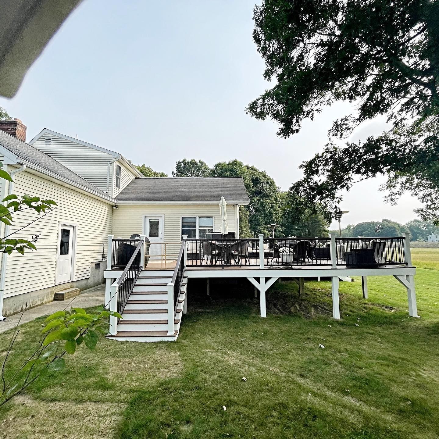 Backyard with a raised wooden deck, stairs, and a house. Green grass and trees surround it.