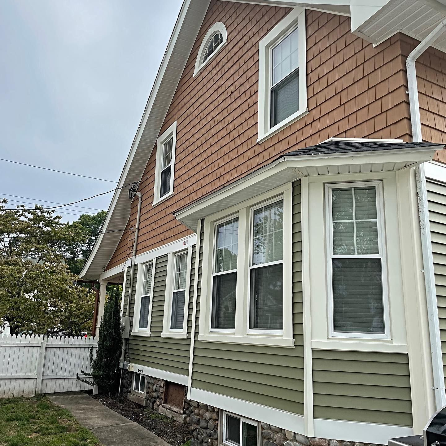 Two-story house with brown shingle siding on top and green siding below, white trim, and a cloudy sky.
