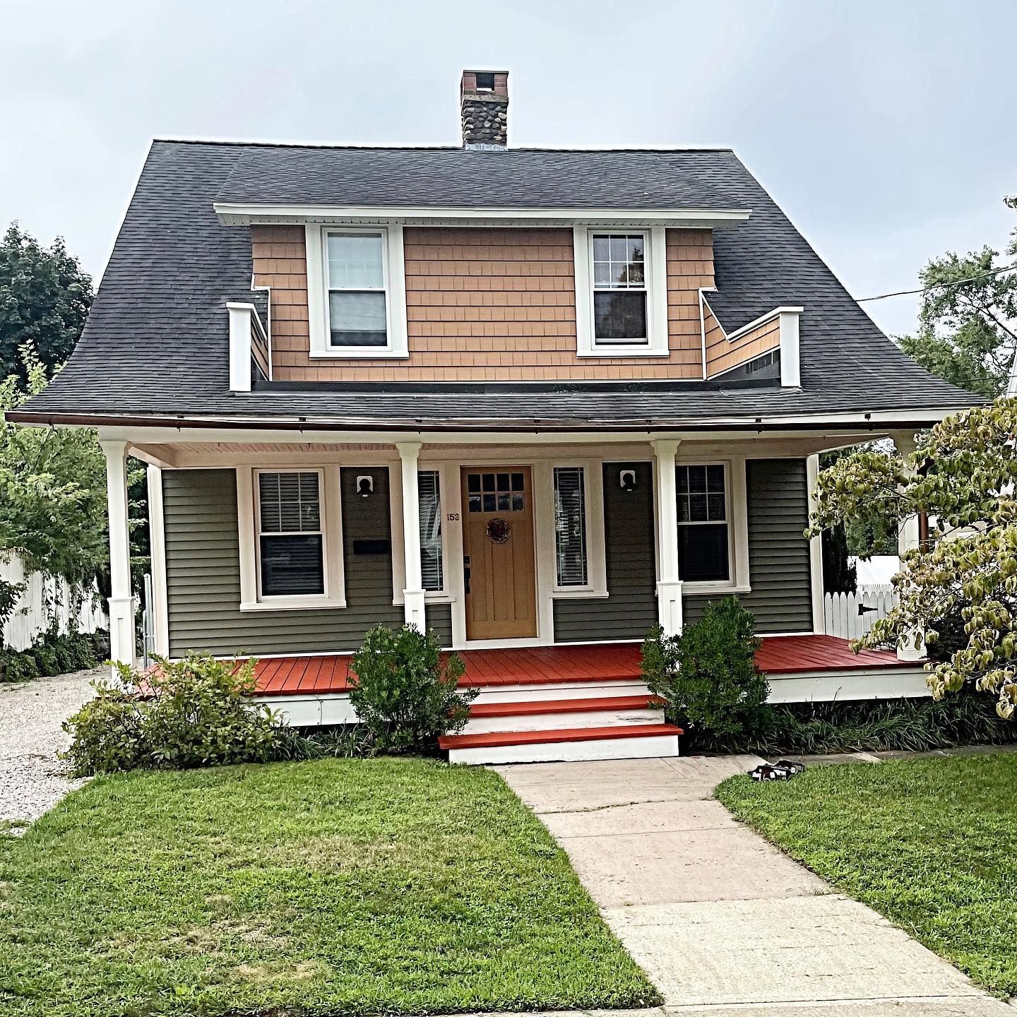 Two-story house with green siding, brown shingle dormer, and porch with red steps, in front of a green lawn and sidewalk.