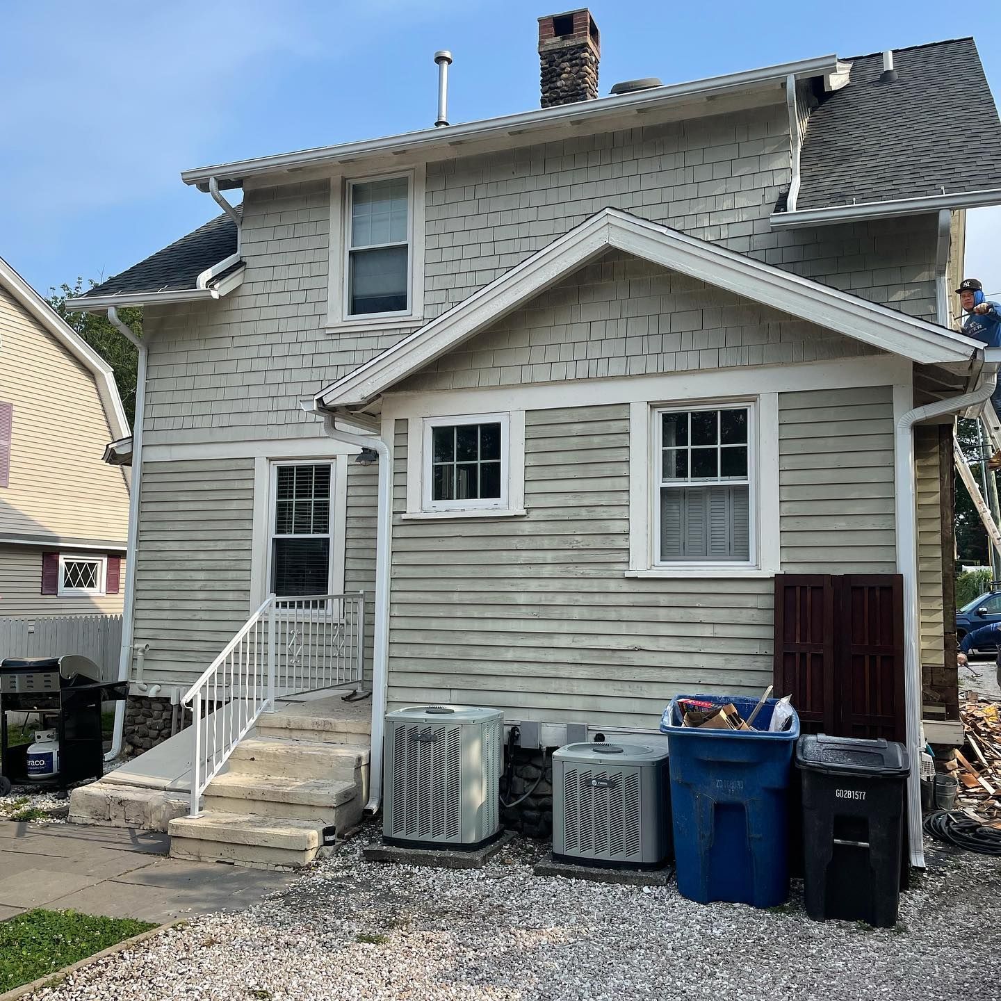 Side view of a two-story house with light-colored siding, two AC units, and trash cans in the yard.