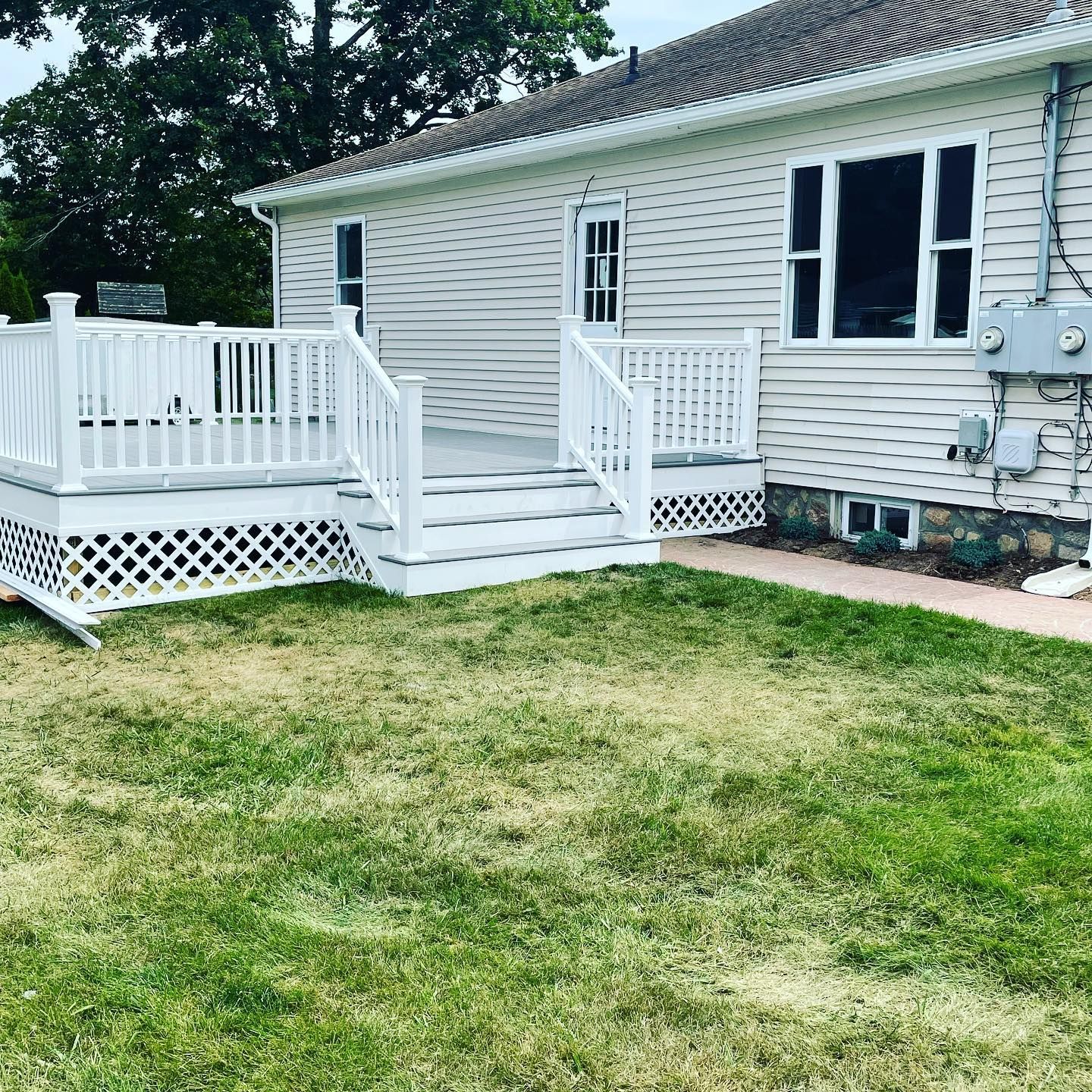 White deck with railings, steps, and latticework attached to a house with beige siding and a patchy green lawn.