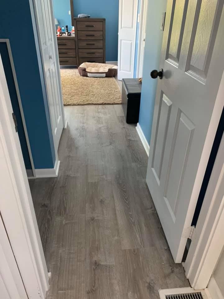 Hallway with gray wood-look flooring, white trim, and blue walls. Bedroom visible at the end.