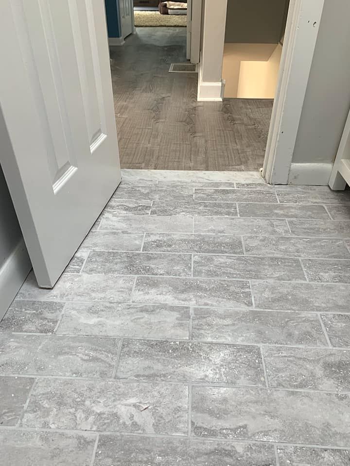 Gray, rectangular tile floor leading into a hallway with a slightly darker floor. A white door is partially open.