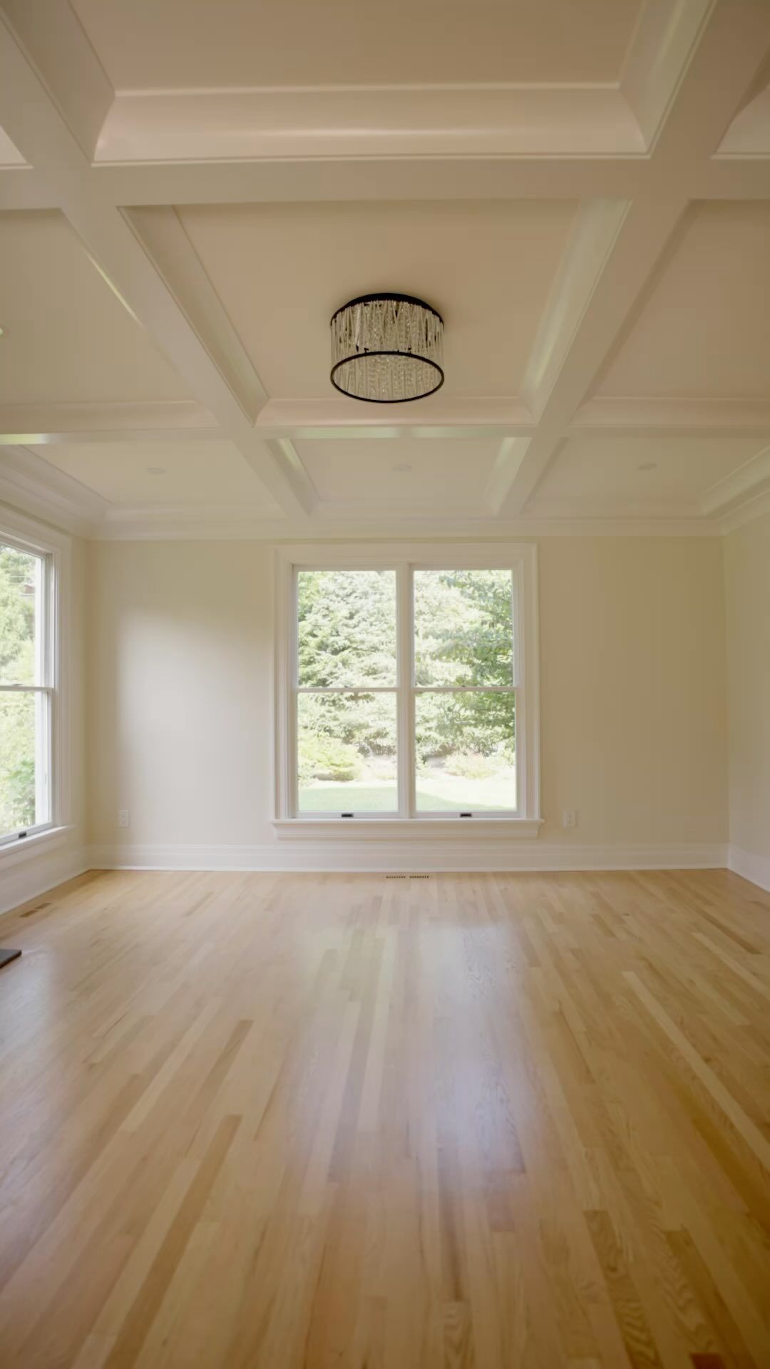 Empty room with light wood floors, coffered ceiling, windows, and chandelier.