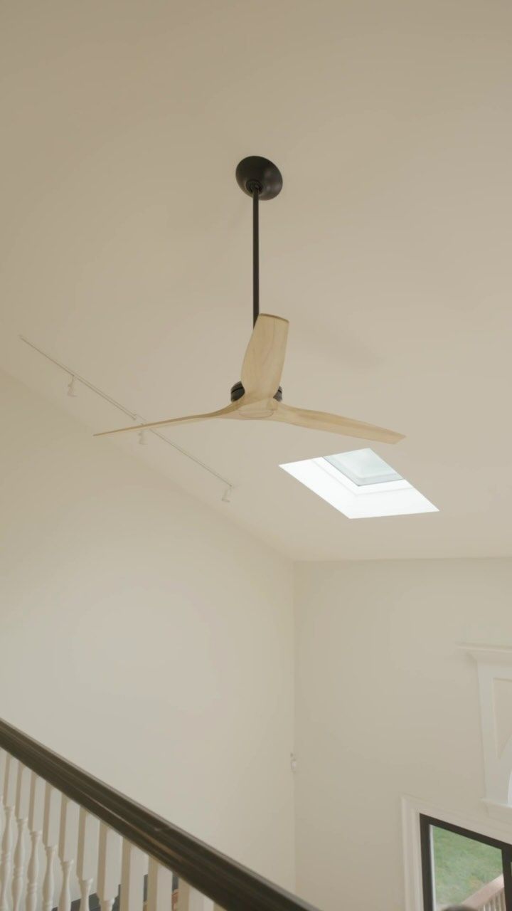 Ceiling fan with wooden blades, black downrod, mounted on a white ceiling above a staircase.