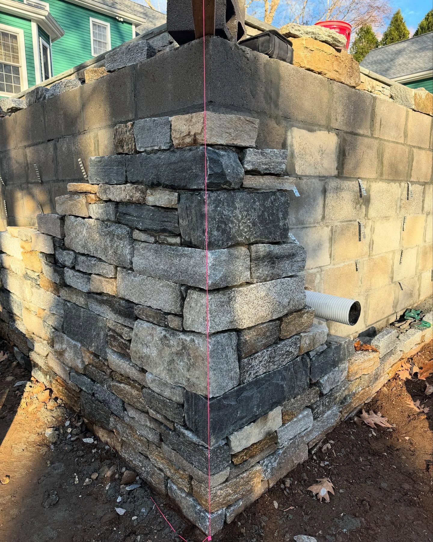 Corner of a stone-veneered wall being constructed; stones are gray, tan, and black. A level guide string is visible.