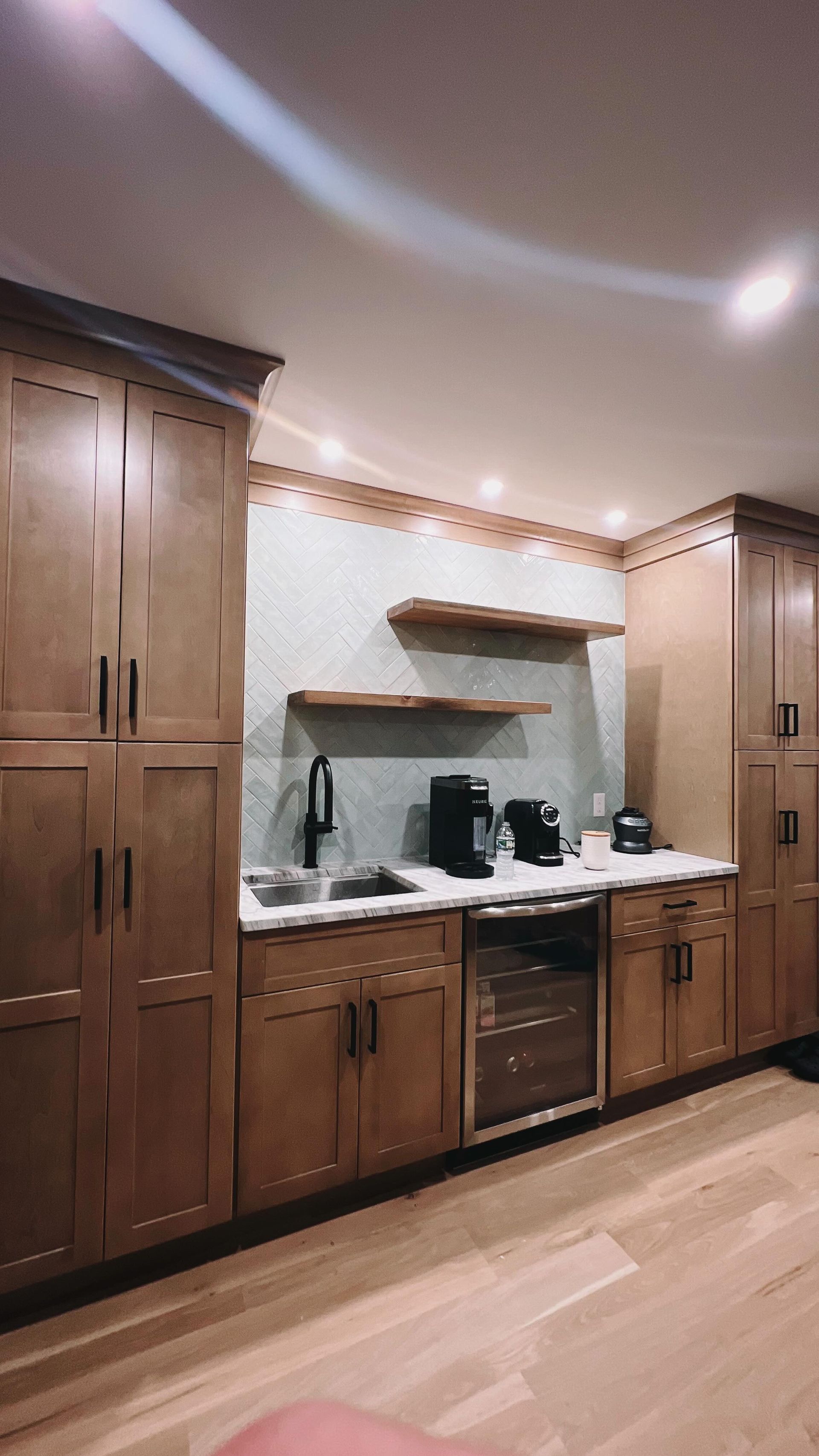 Wooden kitchen cabinets with a coffee station, light-colored countertops, and two wooden shelves against a textured backsplash.