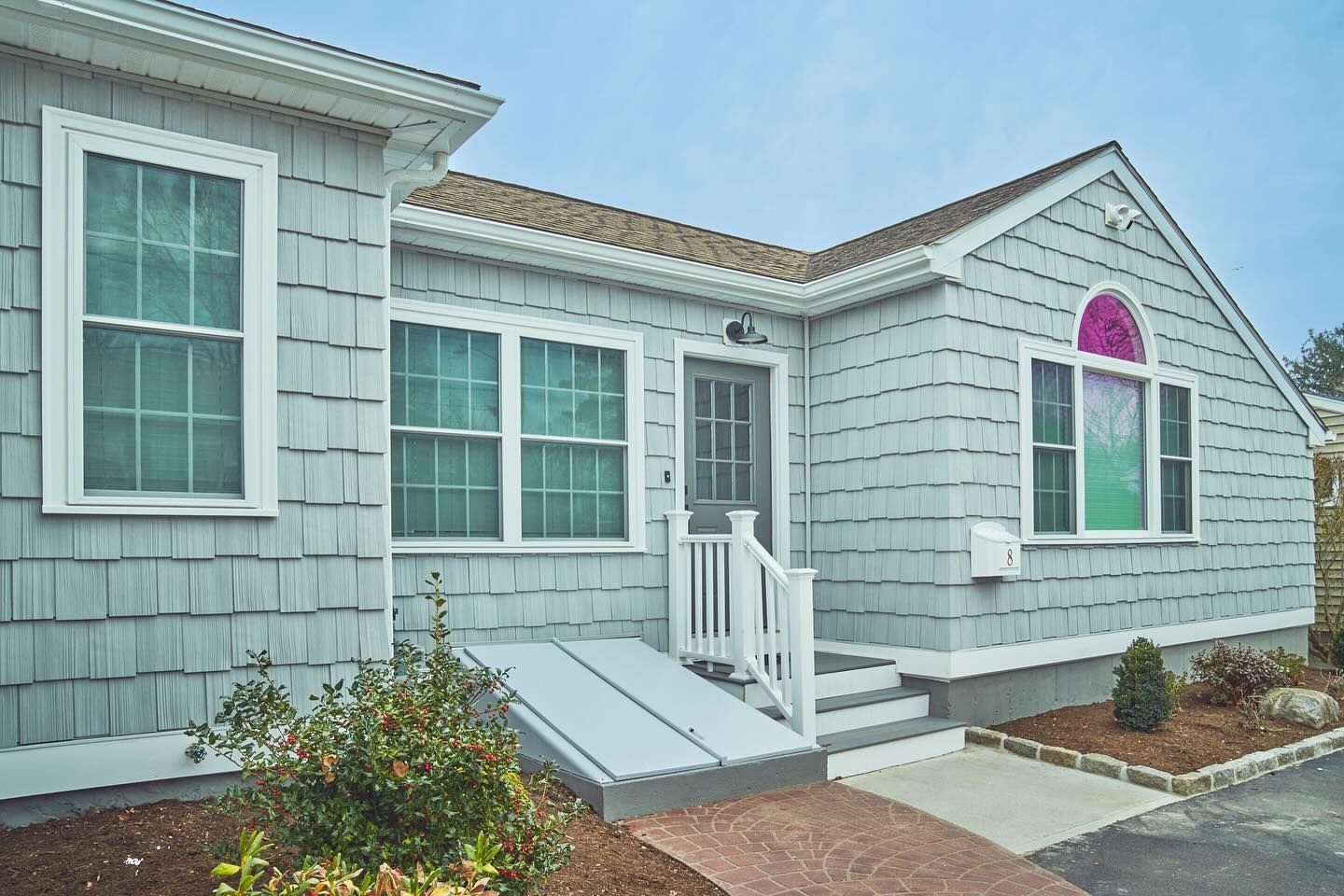 A light gray house with white trim, stairs, and a ramp. Brown roof, green bushes, and a pink-arched window.