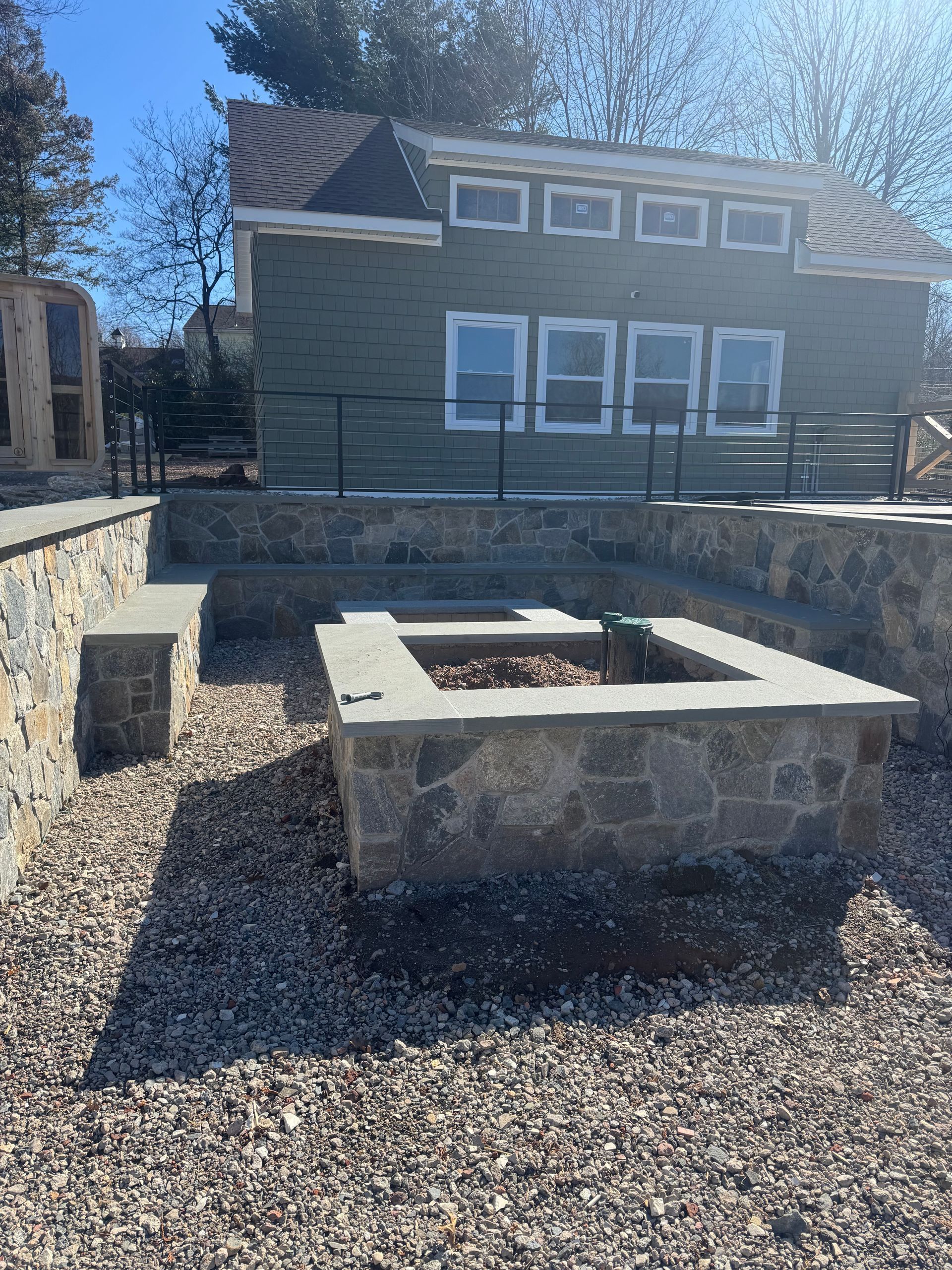 Stone fire pit and seating area in front of a green building with white-framed windows, and gravel ground.