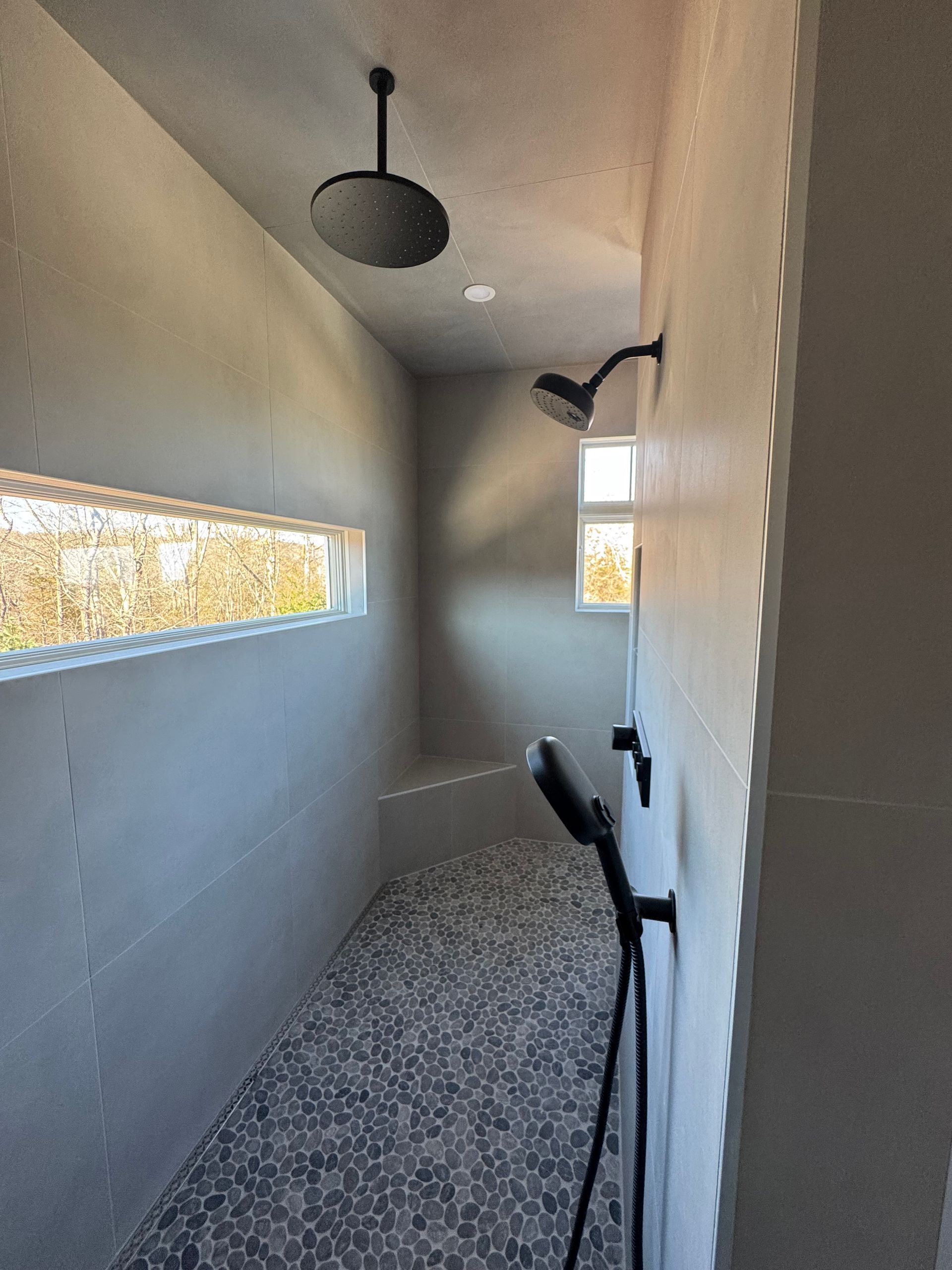 Long, narrow modern shower with pebble floor, gray walls, two shower heads, and a window.
