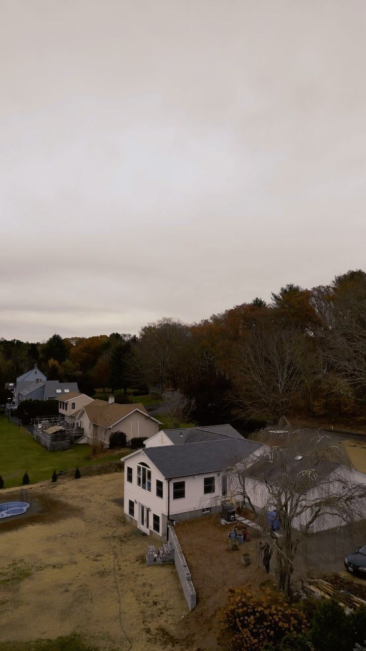Houses and trees under a cloudy sky. Brown grass in the foreground and a small pool on the left.