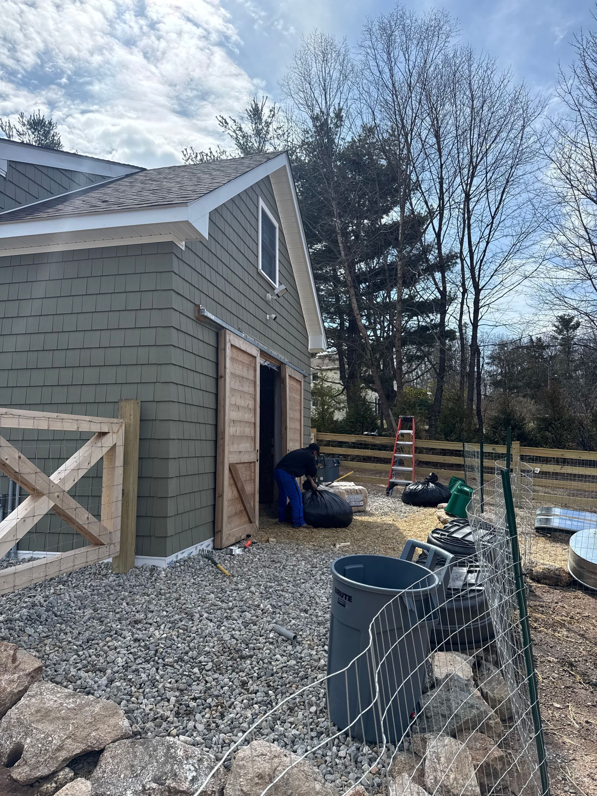 A person is filling a feed bucket near a green-sided barn with wooden doors, a fence, and gravel.