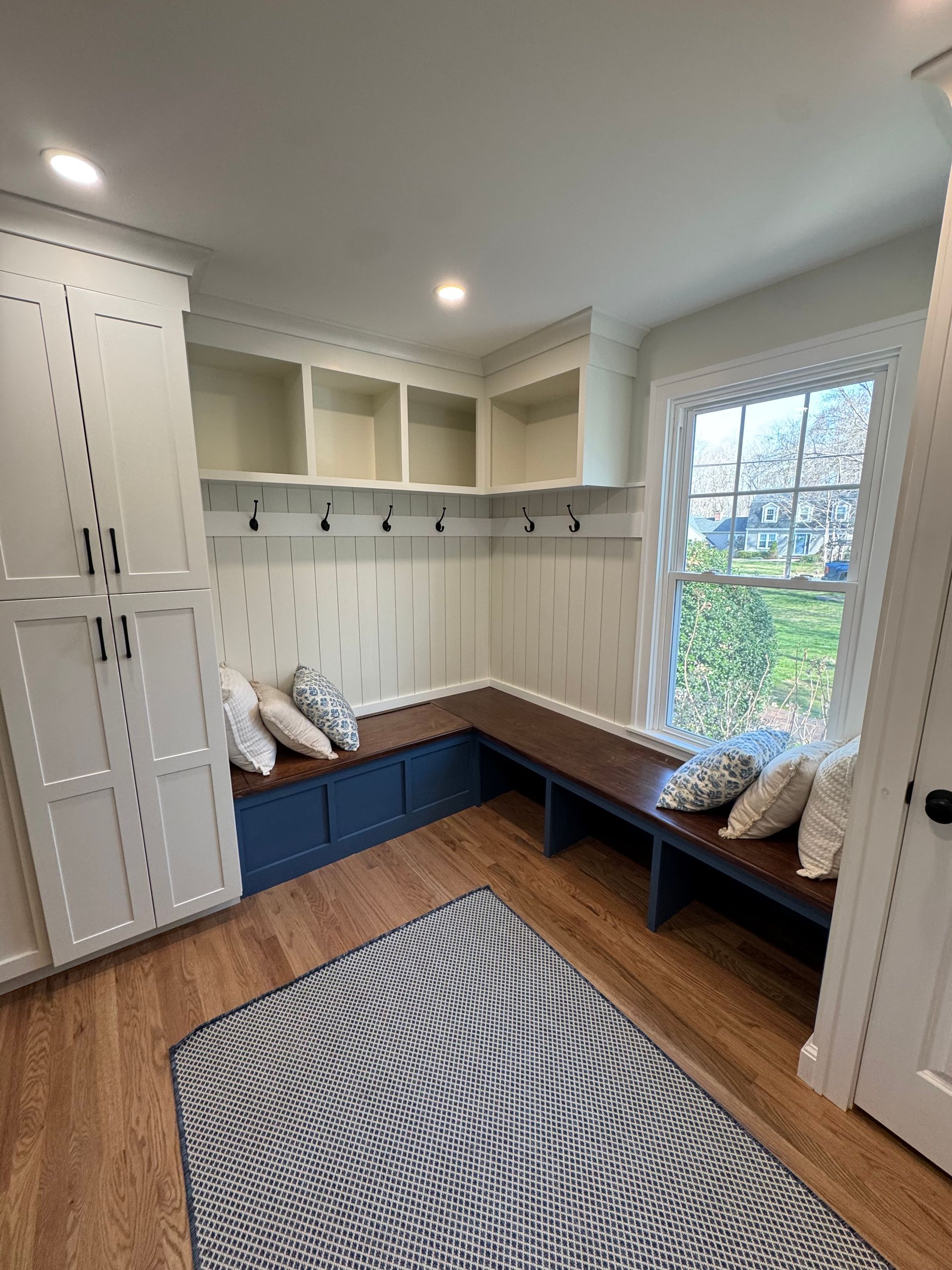 Mudroom with built-in bench, storage cabinets, coat hooks, and area rug. White and blue accents, wood floors, and window.