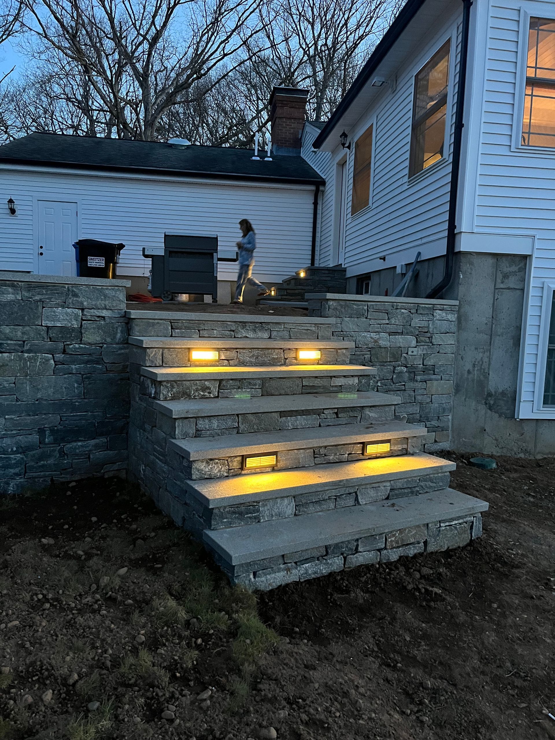 Stone steps with built-in lights lead up to a white house; a person stands at the top near a detached garage.