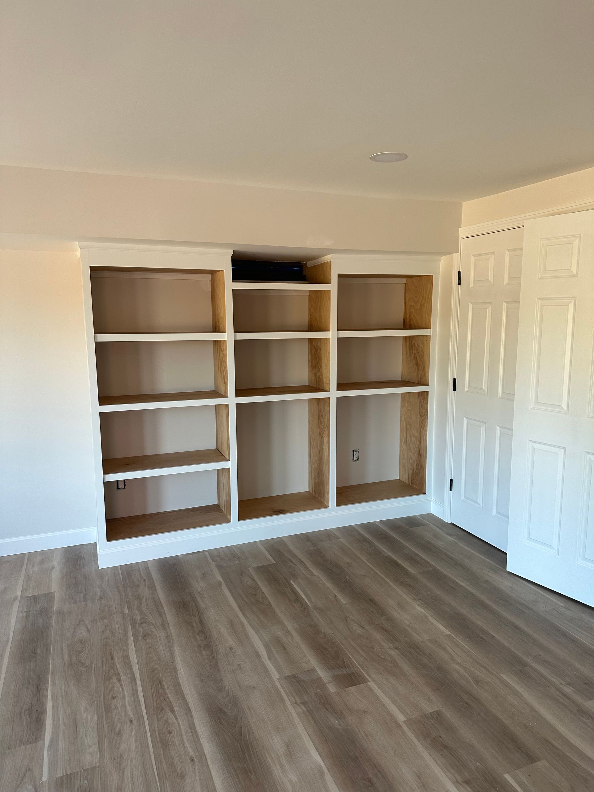 White built-in bookshelves with wooden shelves in a room with light wood-look flooring and a white door.