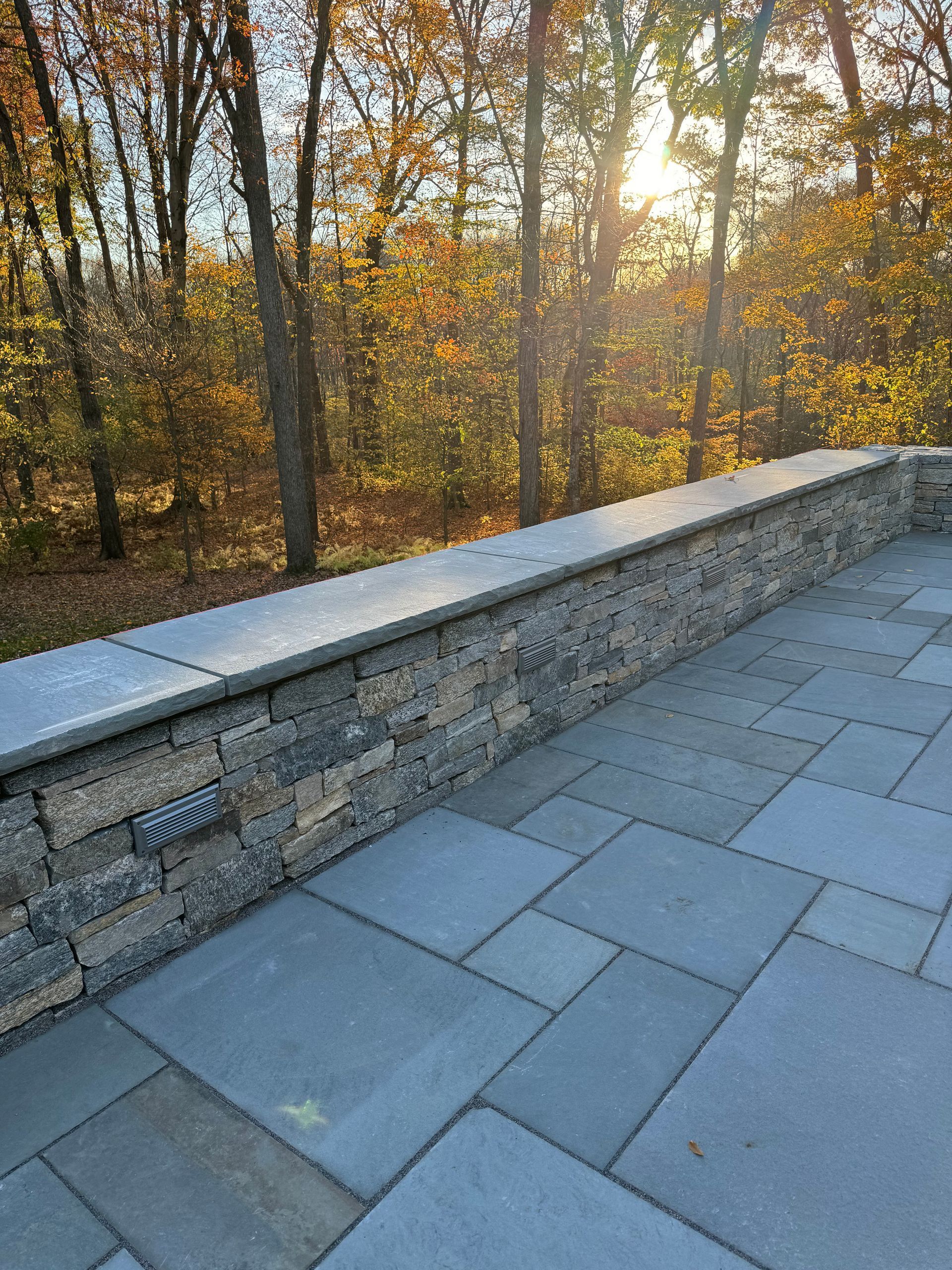 Stone patio with a low stone wall and trees in the background, lit by the sun.