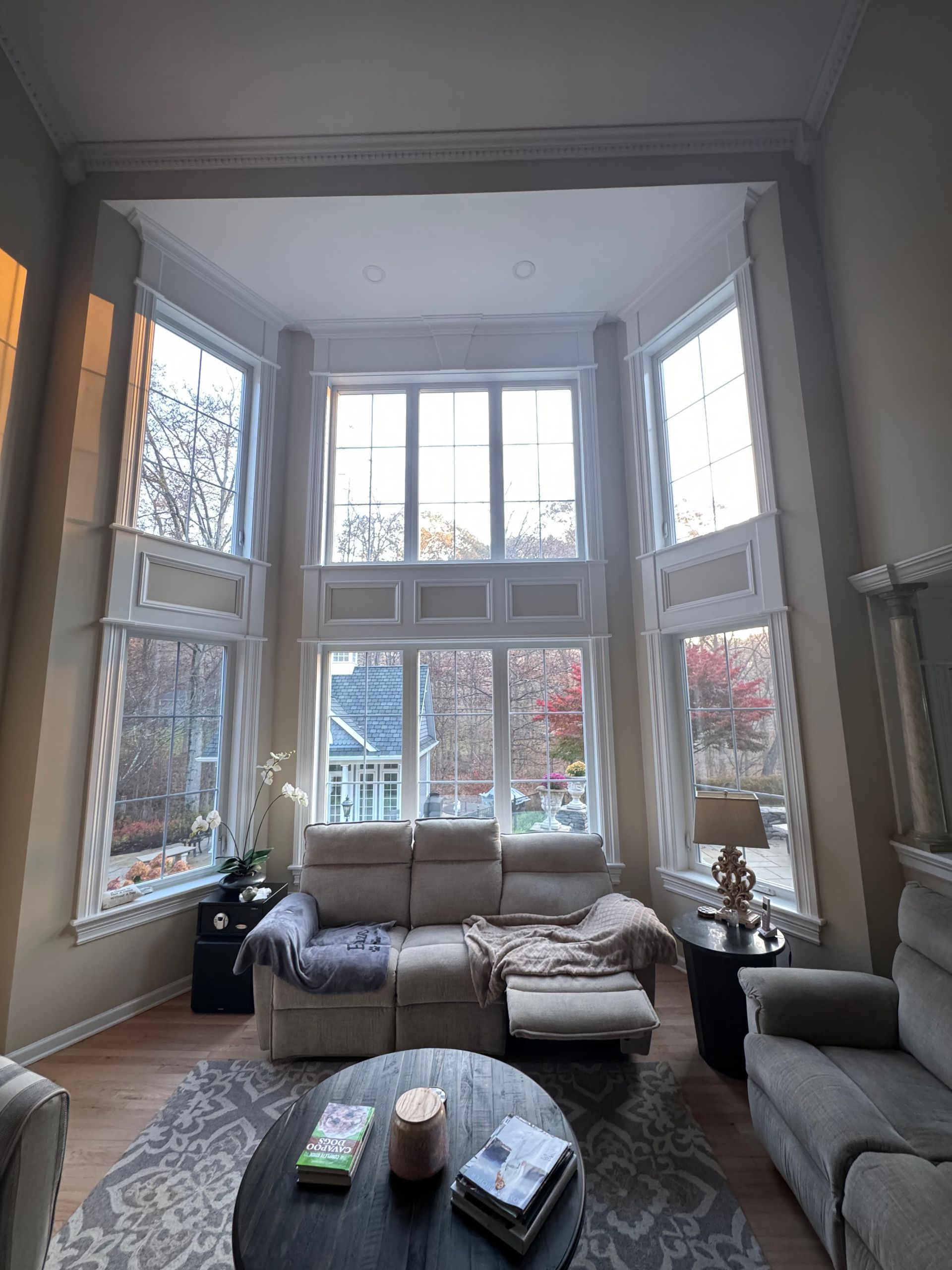 Living room with large bay windows, beige couches, wooden floor, and a round coffee table.