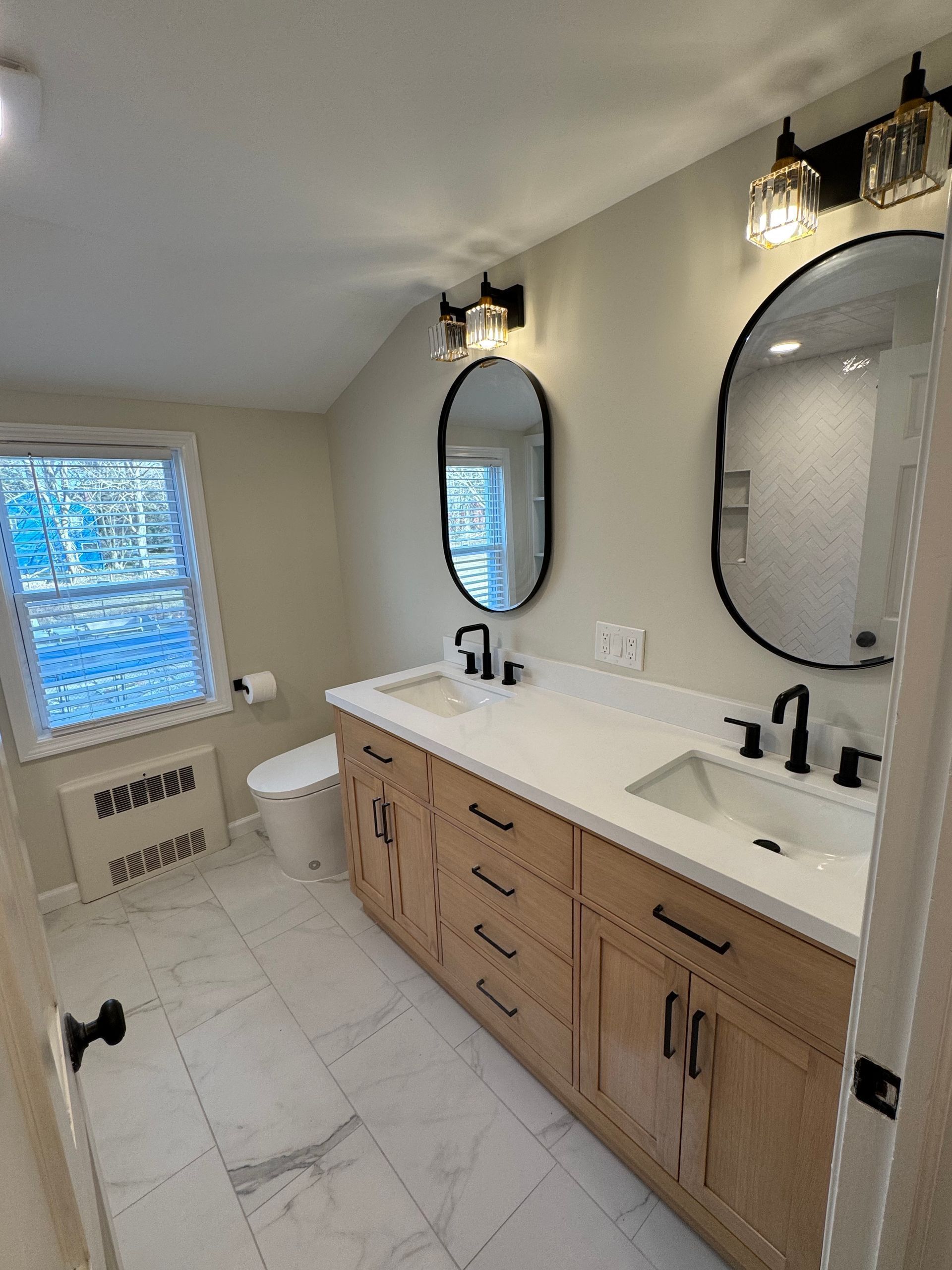 Bathroom with light wood vanity, white countertops, two black-framed oval mirrors, and marble flooring.