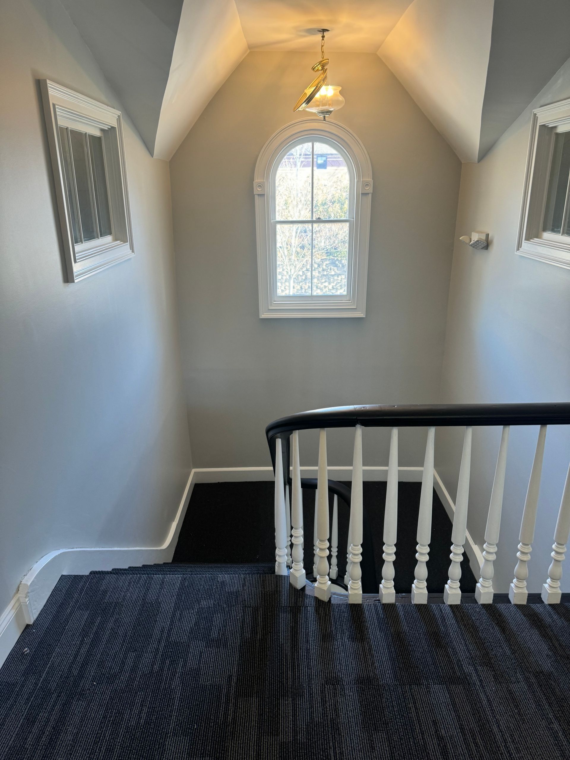 Staircase with gray walls, black carpet, white banister, arched window at top.
