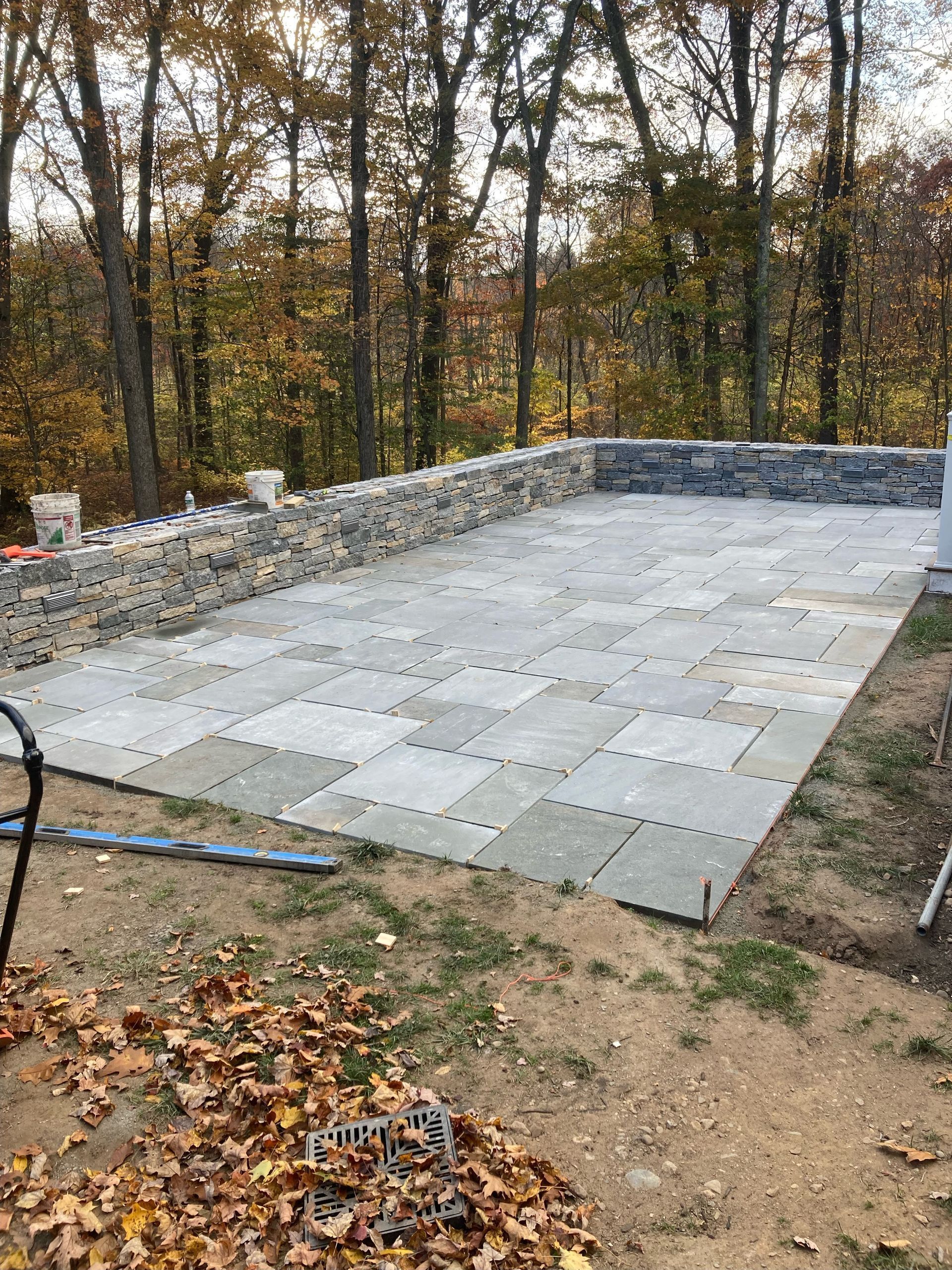 Stone patio under construction with stone retaining wall in a wooded area.