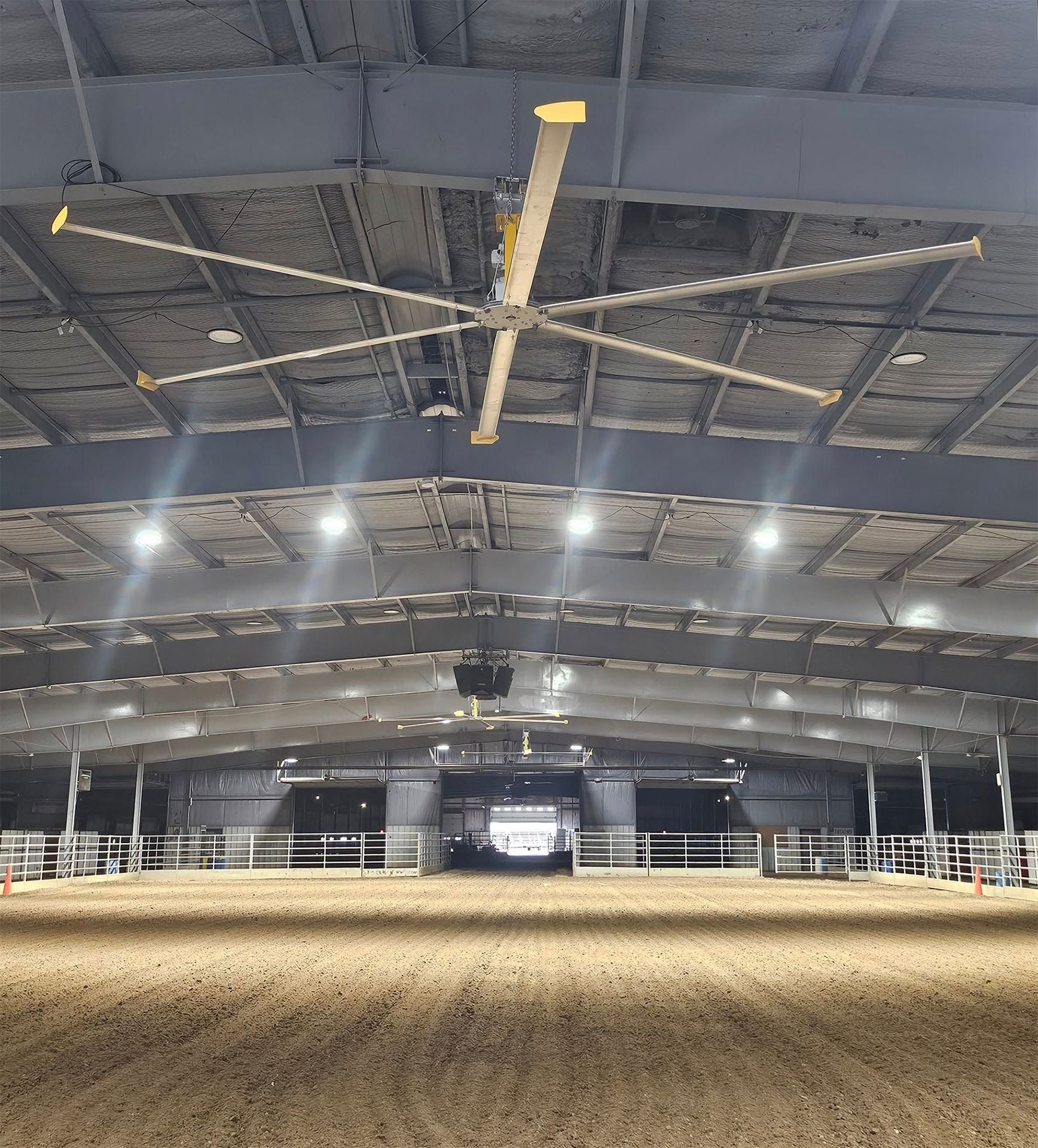 Large indoor riding arena with dirt floor, high ceiling, and industrial fan.