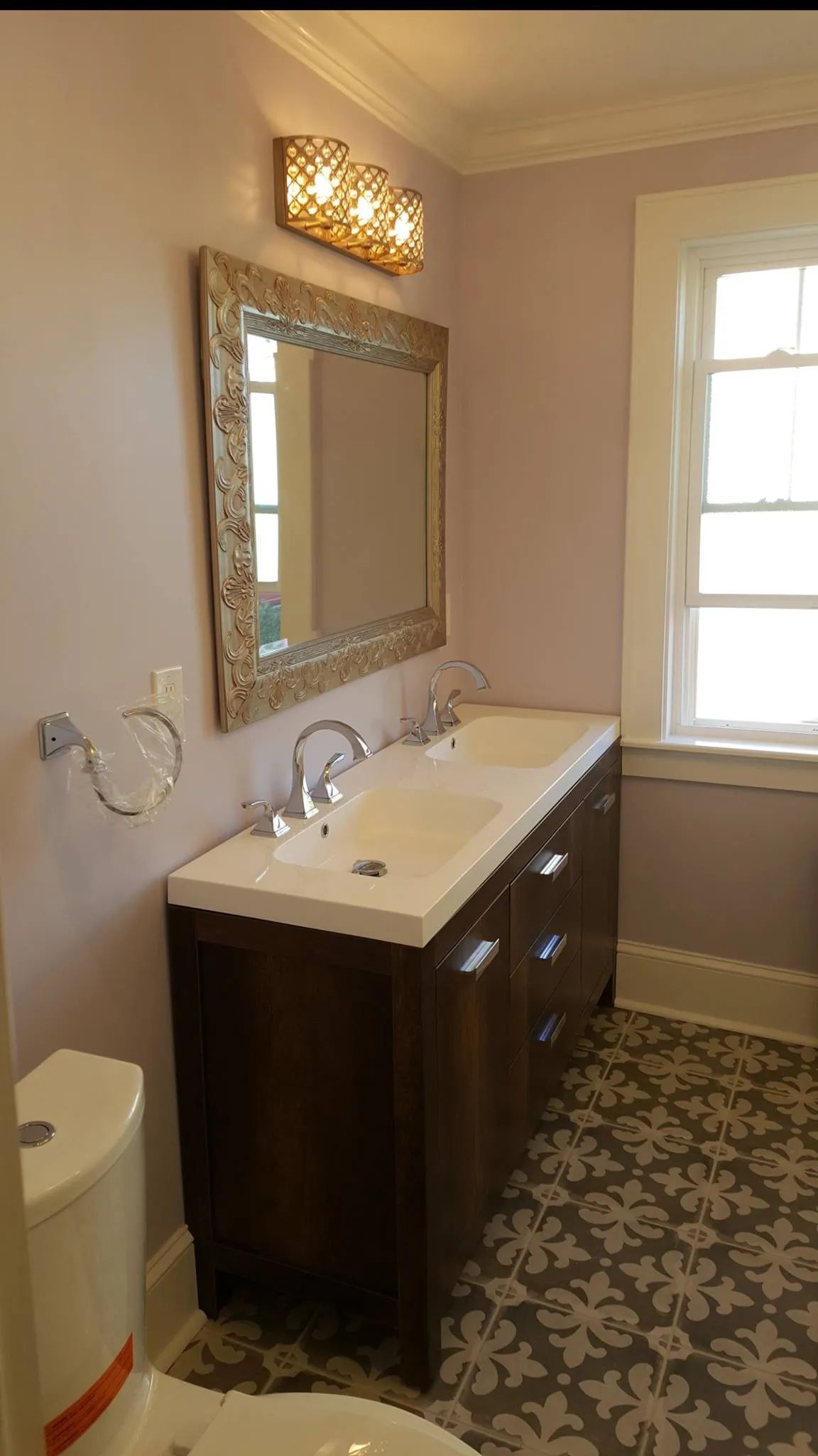 Small bathroom with dark vanity, gold mirror, floral floor, and window.