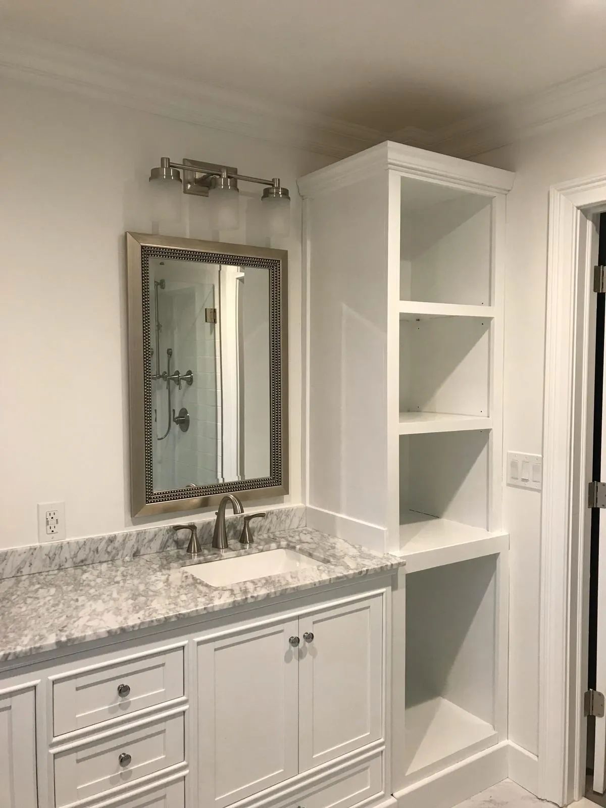 Bathroom vanity with marble countertop, mirror, and white built-in shelving cabinet.