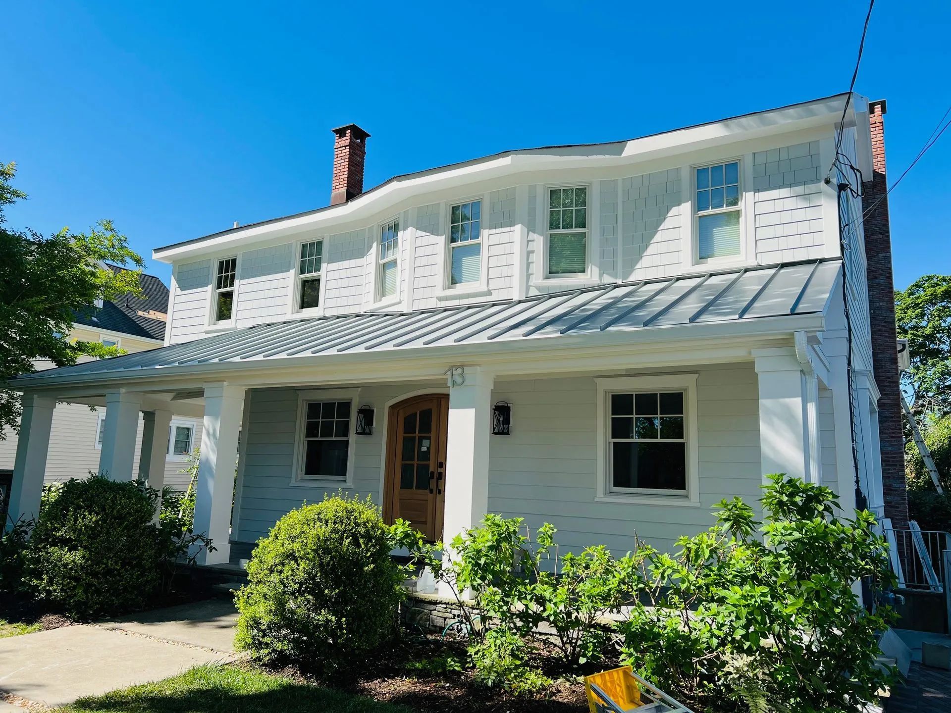 White two-story house with a front porch, blue shutters, and bushes under a clear sky
