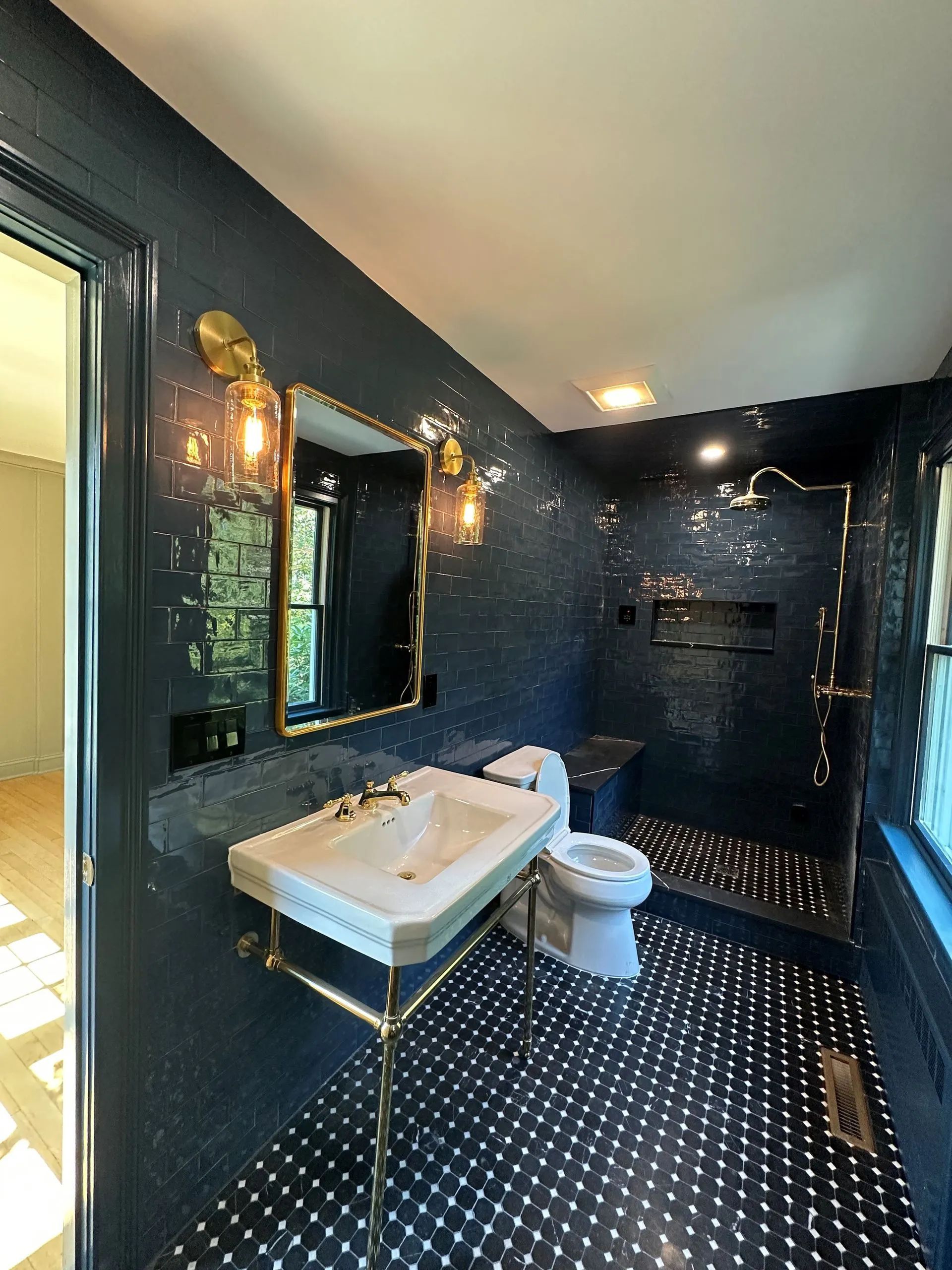 Dark blue tiled bathroom with a sink, toilet, shower area, and brass wall sconces