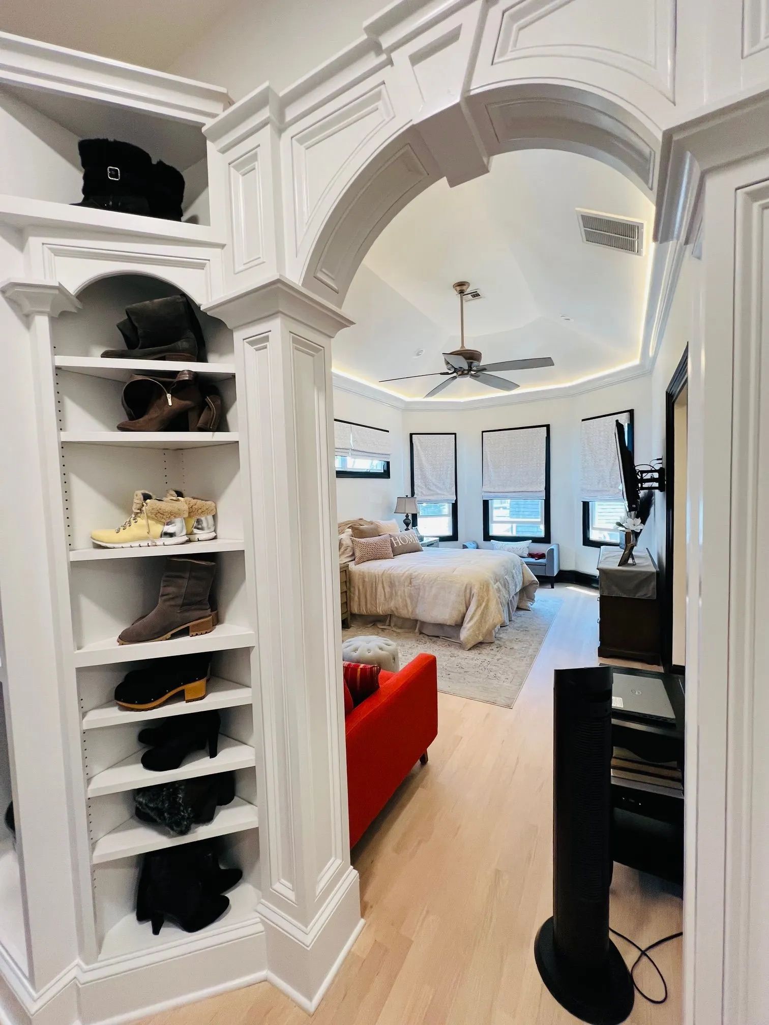 Bright white bedroom viewed through an ornate archway, with shelves on the left and a bed, windows, and fan inside.