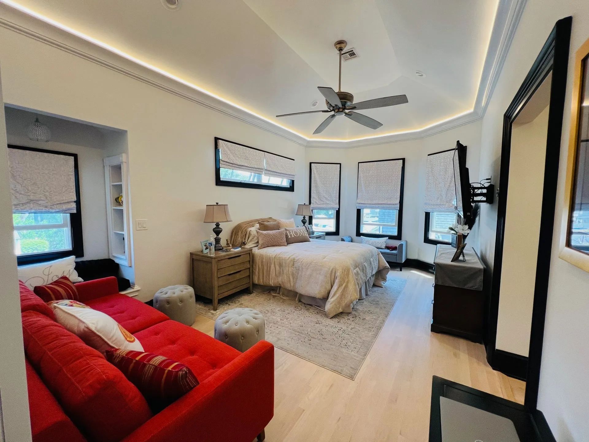 Bright bedroom with a red couch, beige bed, ceiling fan, and large windows with natural light.