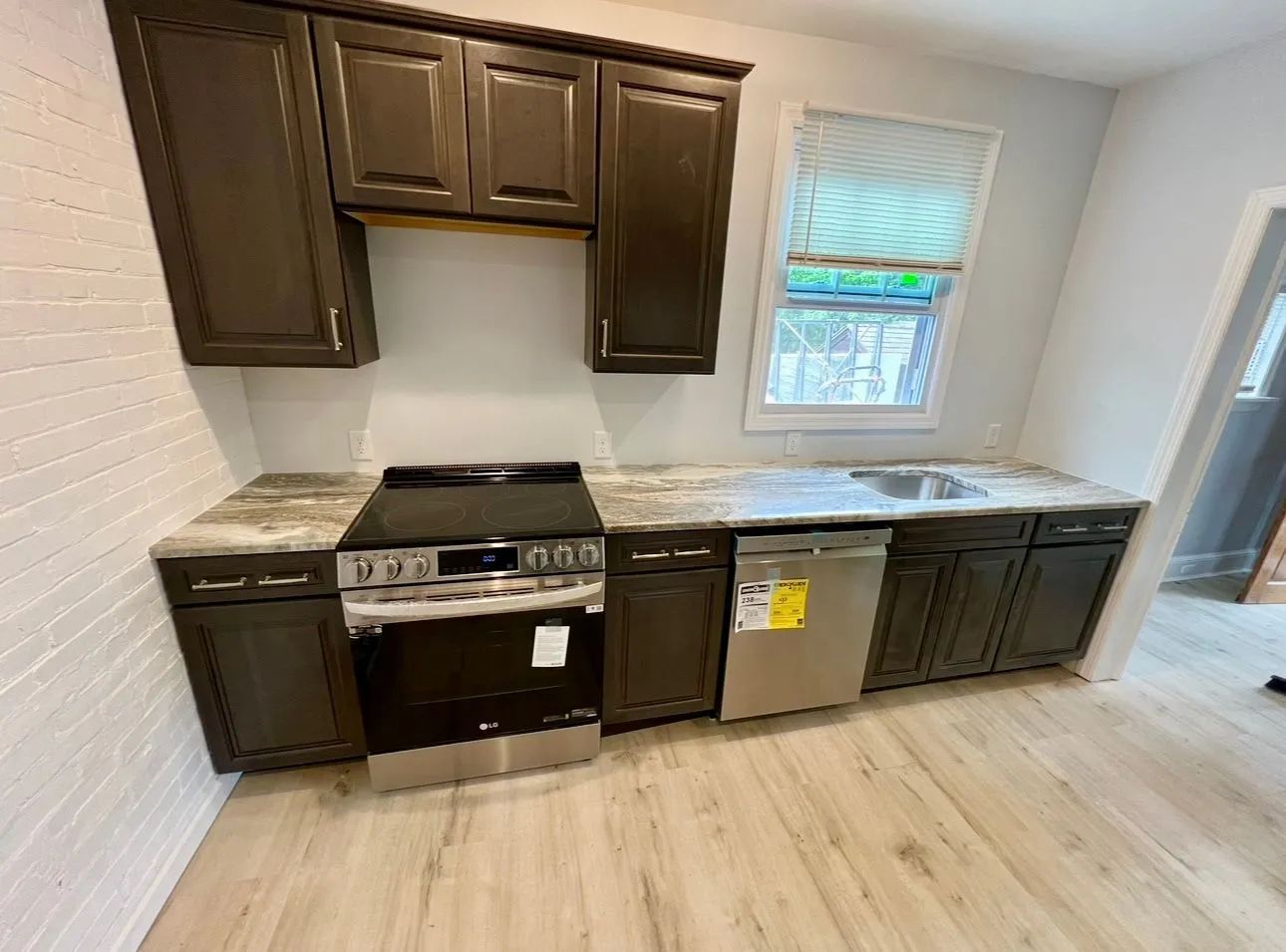 Small kitchen with dark cabinets, stove, countertop, sink, window, and light wood flooring.