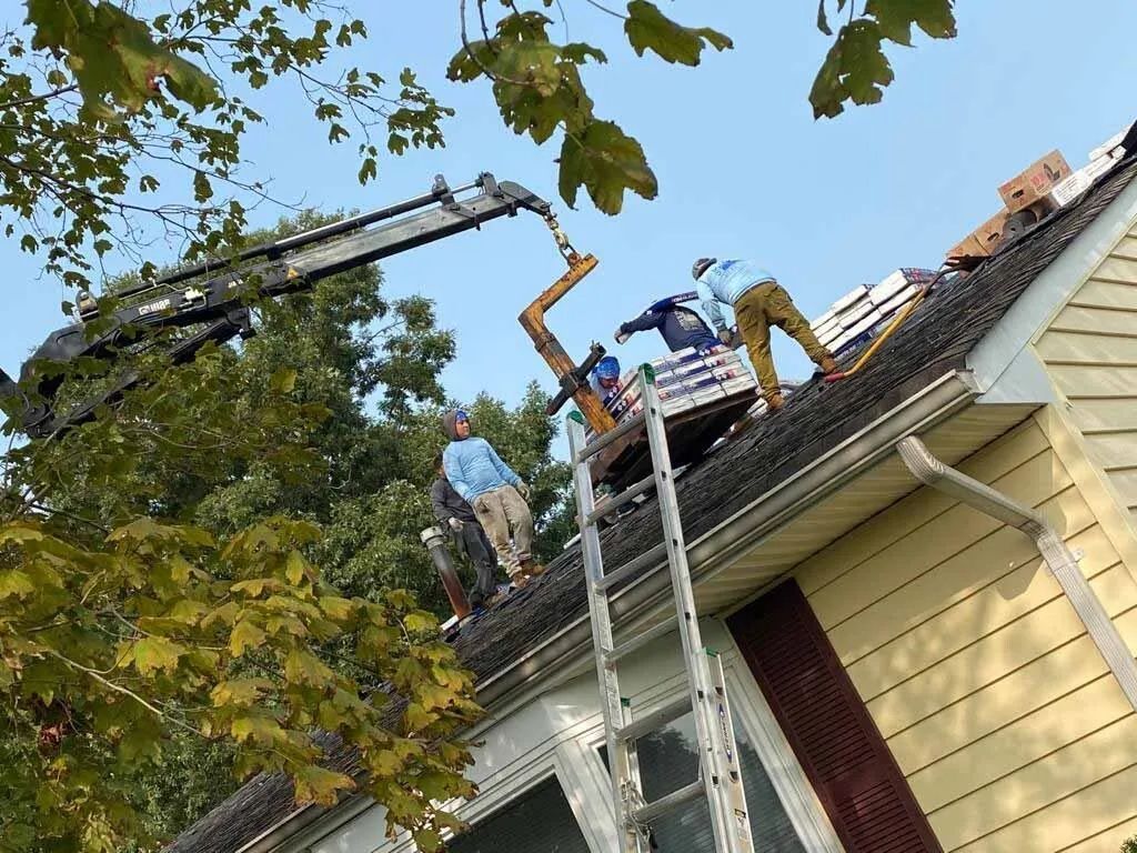 Workers on a roof beside a ladder, using a crane to lift roofing materials near a house and trees.