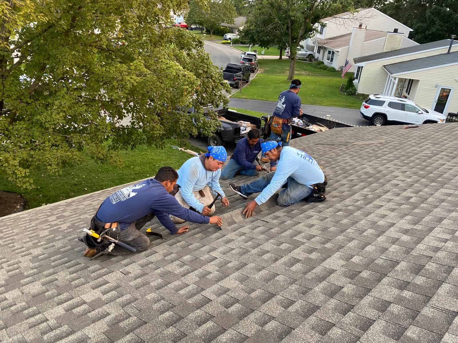Workers repairing a shingled roof on a house, kneeling in a line near the ridge.